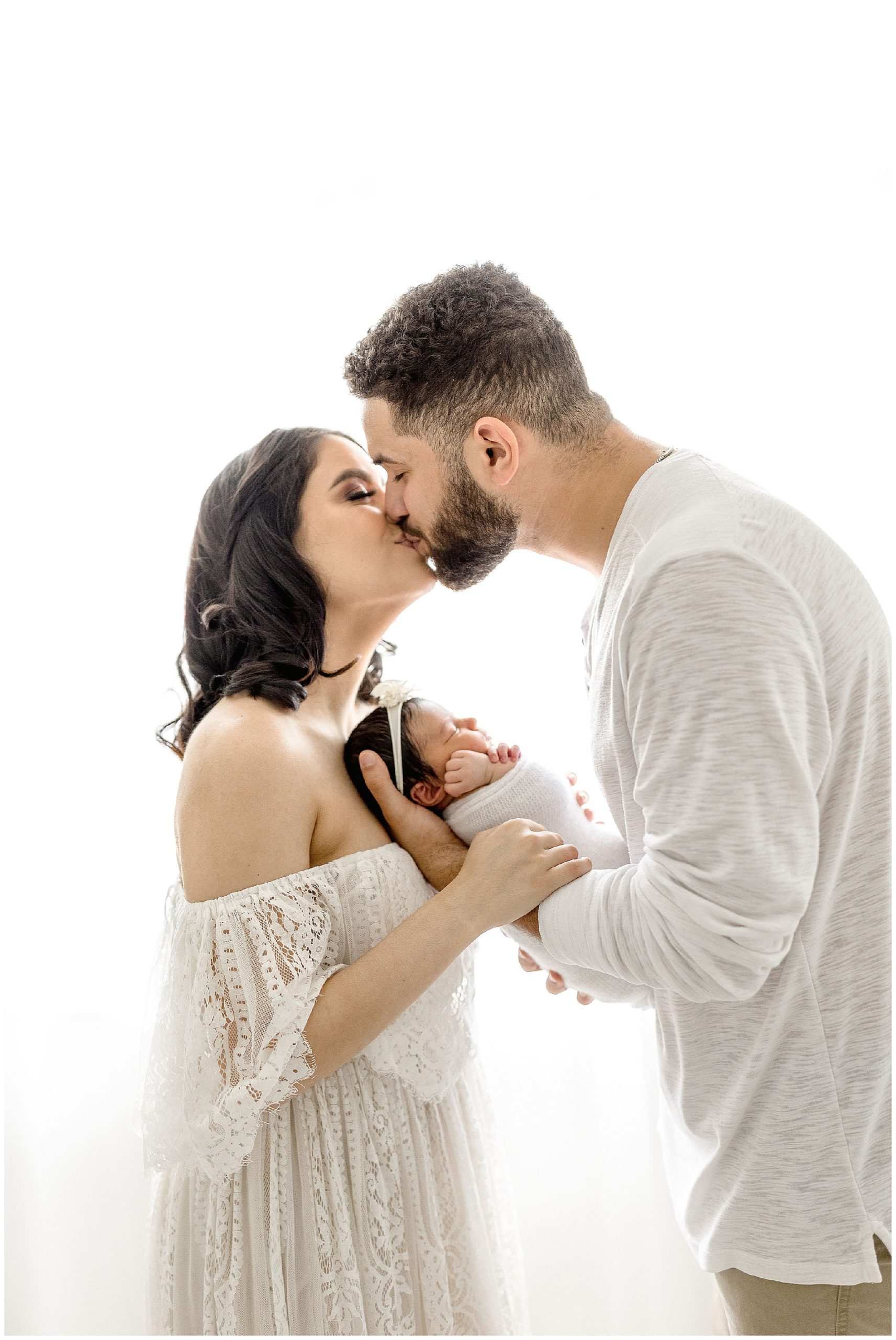 New parents kiss while holding their newborn. Photo by Ivanna Vidal Photography.