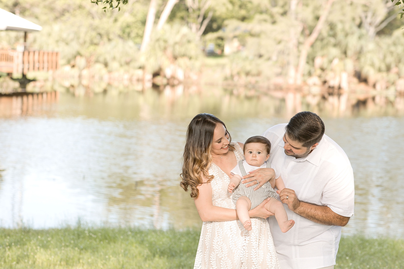 Mom and dad embrace baby boy near river with Miami Children Photographer. Photo by Ivanna Vidal Photography.