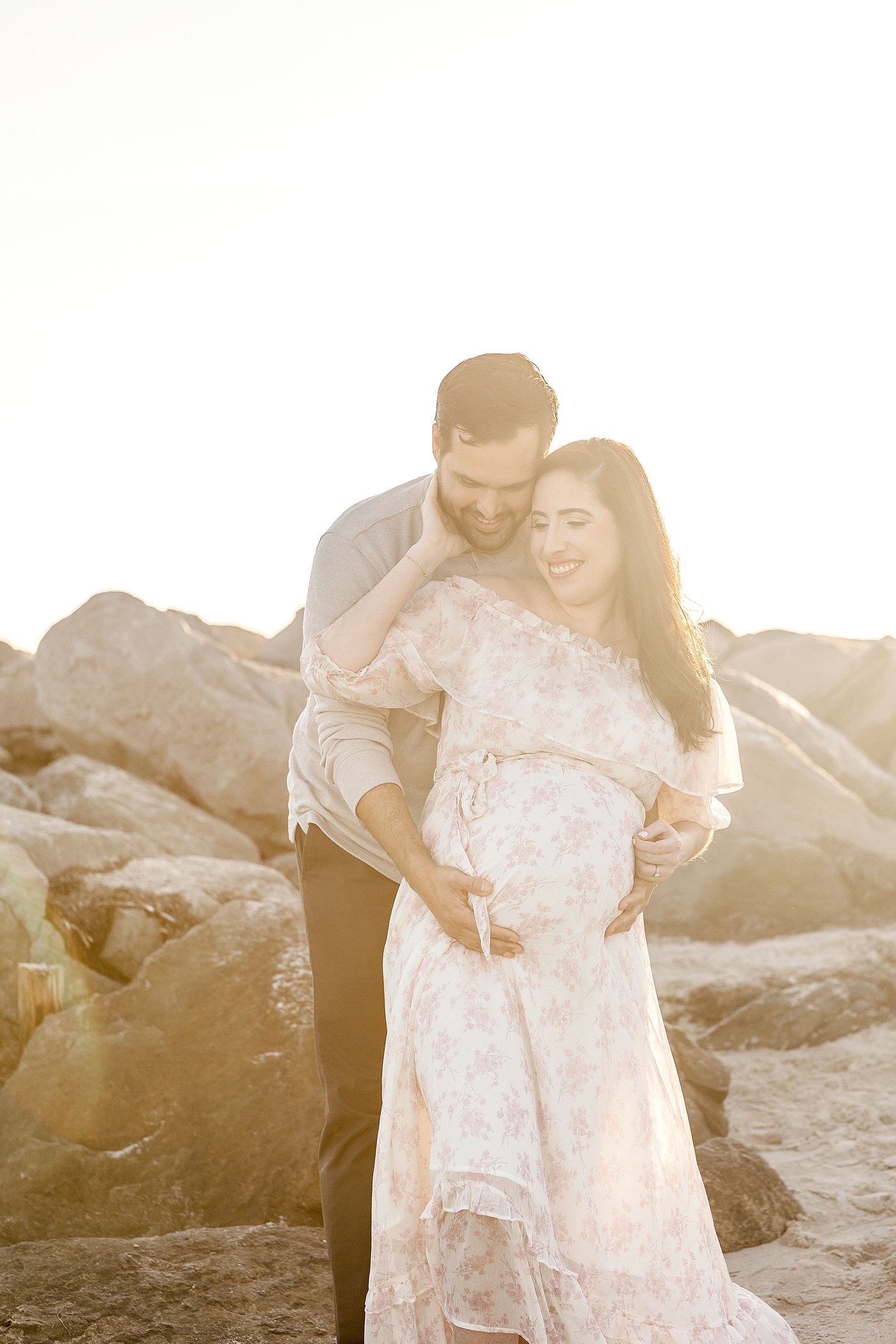 Expectant parents to be embrace one another in front of rocks during maternity session at El Farito Beach Miami FL. Photo by Ivanna Vidal Photography.