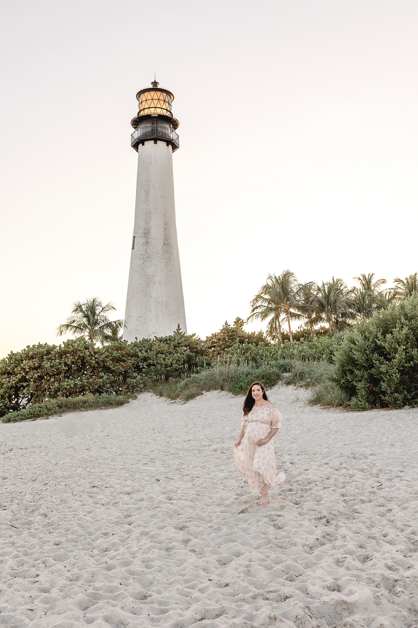 Mom to be in white dress stands in front of El Farito Beach Miami FL. Photo by Ivanna Vidal Photography.