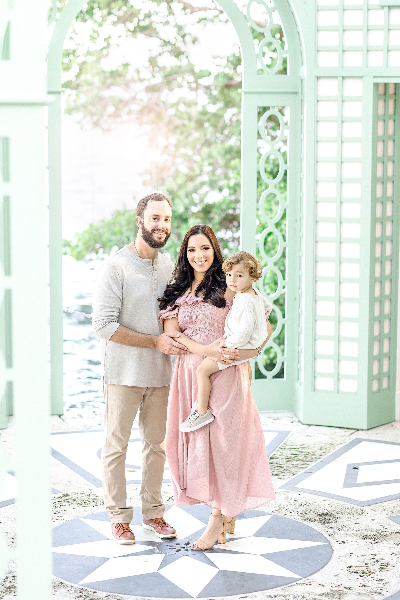 Family of three stands in front of green gate during Miami family photoshoot at Vizcaya. Photo by Ivanna Vidal Photography.