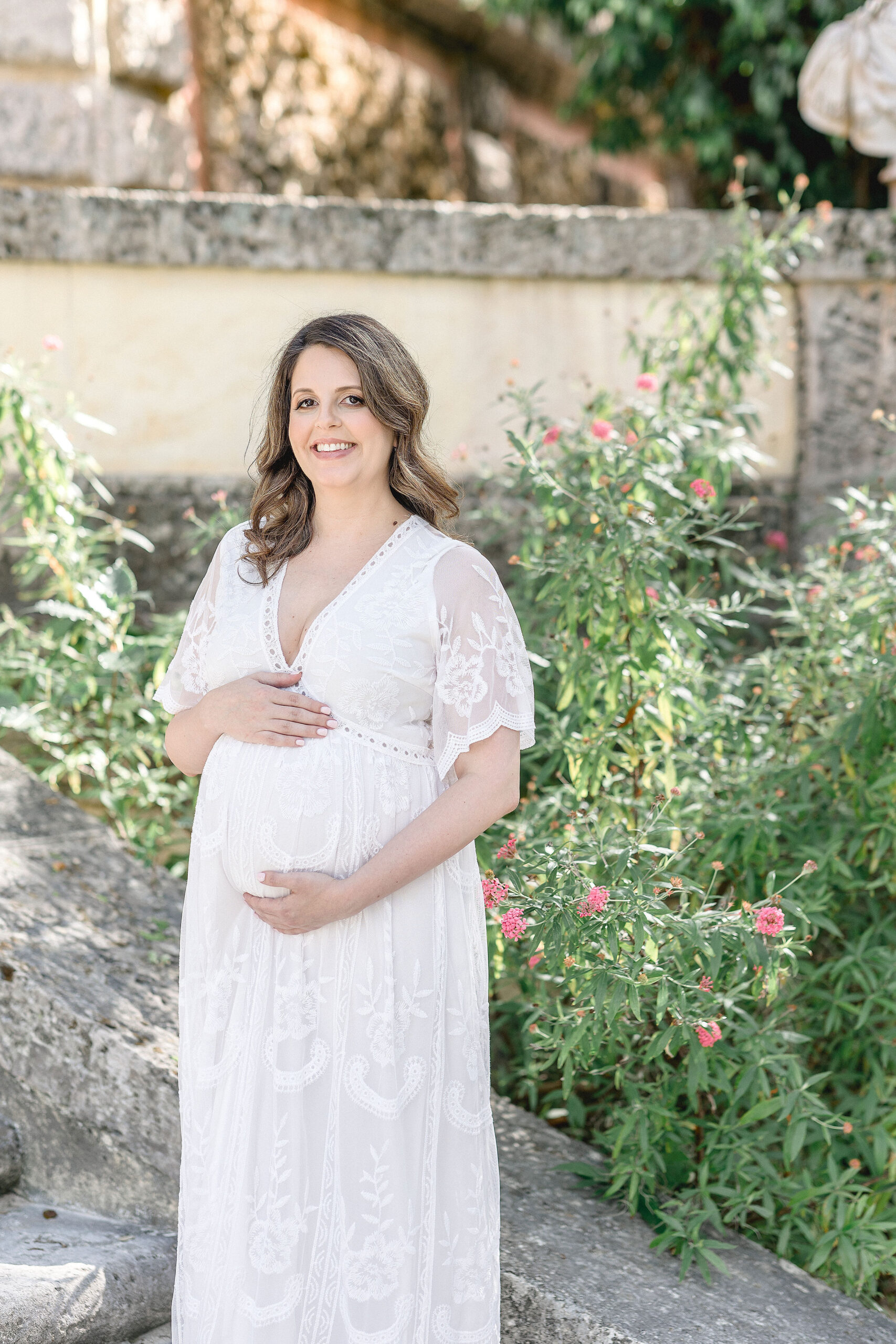 mom to be in white maternity Maternity Clothing Miami standing in a garden holding her bump