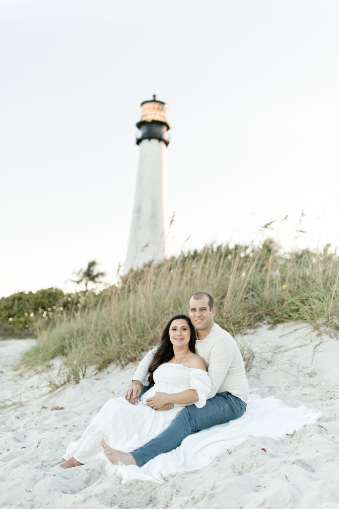 mom and dad to be in white sitting in the sand at Bill Baggs Lighthouse Maternity Photoshoot locations in Miami
