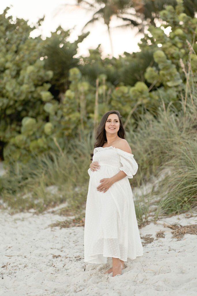 pregnant woman in white maternity gown cradling her bump on the beach Magnolia Birth House