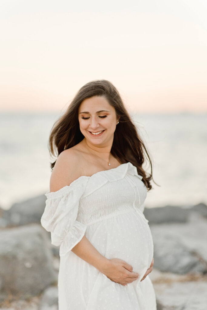 mom to be in white maternity gown looking towards her bump on the beach
