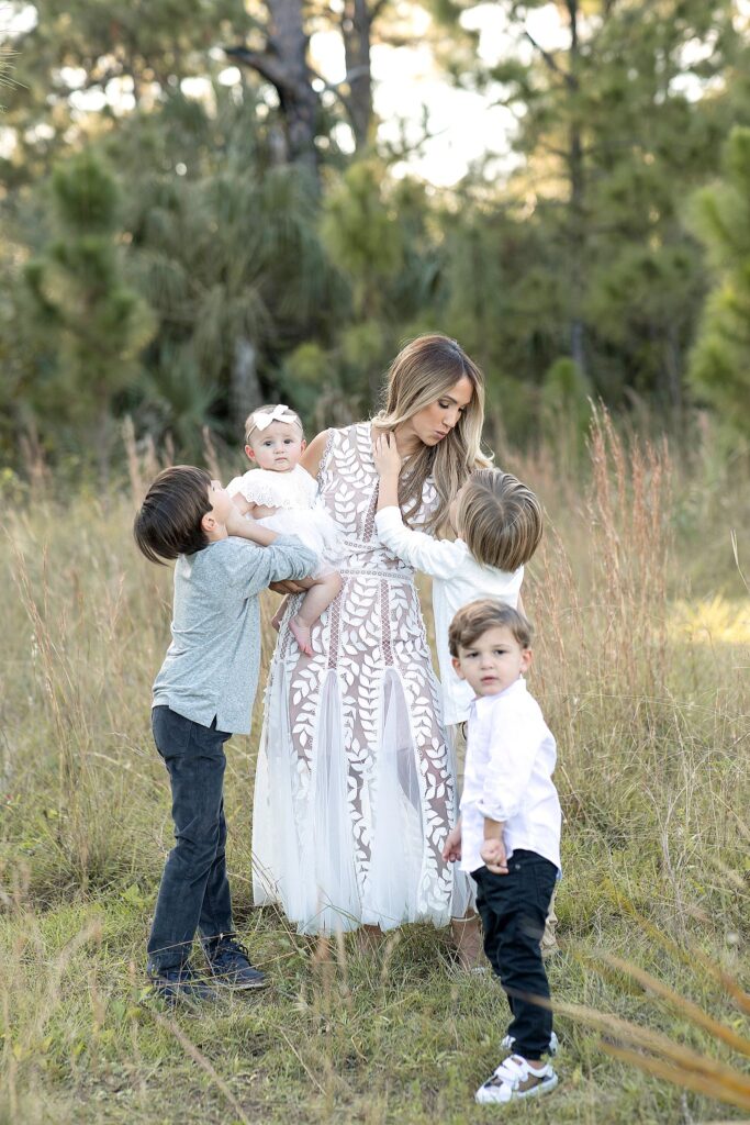 Mom and her three older sons play with her one year old daughter in a grassy field on the edge of the woods