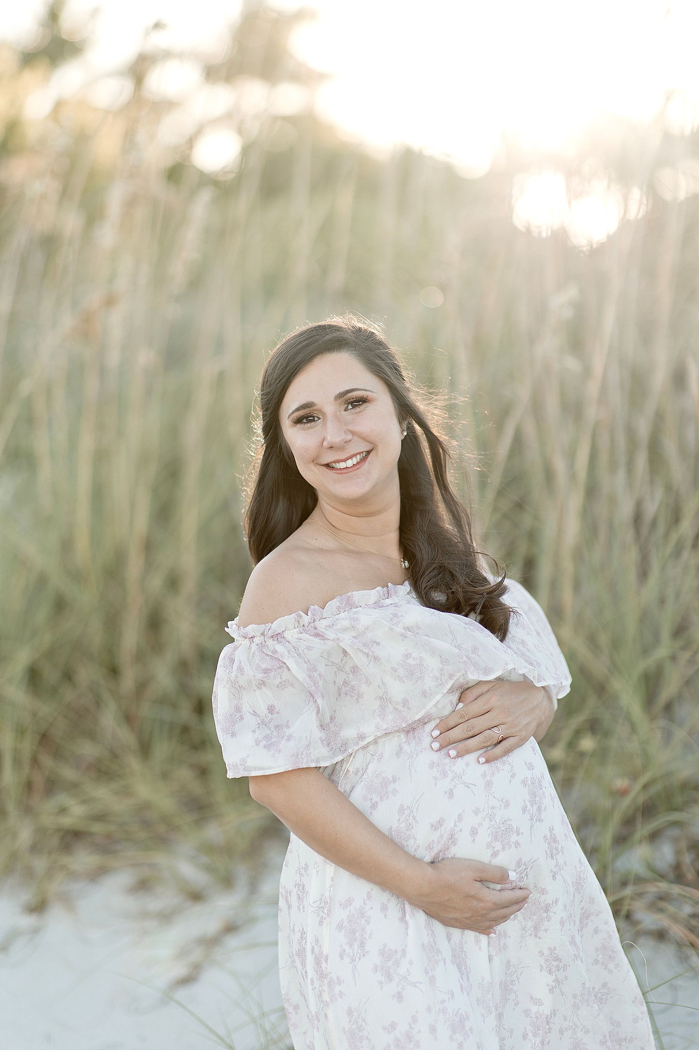 mom to be in white maternity dress cradling her bump on the beach