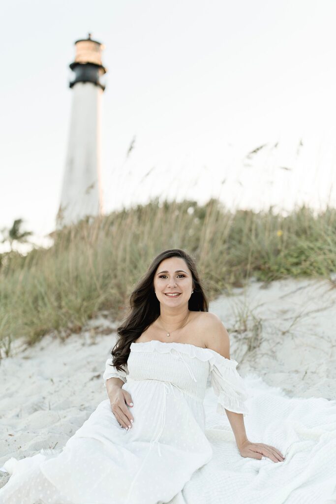 mom to be in white gown sitting in the sand with a lighthouse in the background Miami water birth