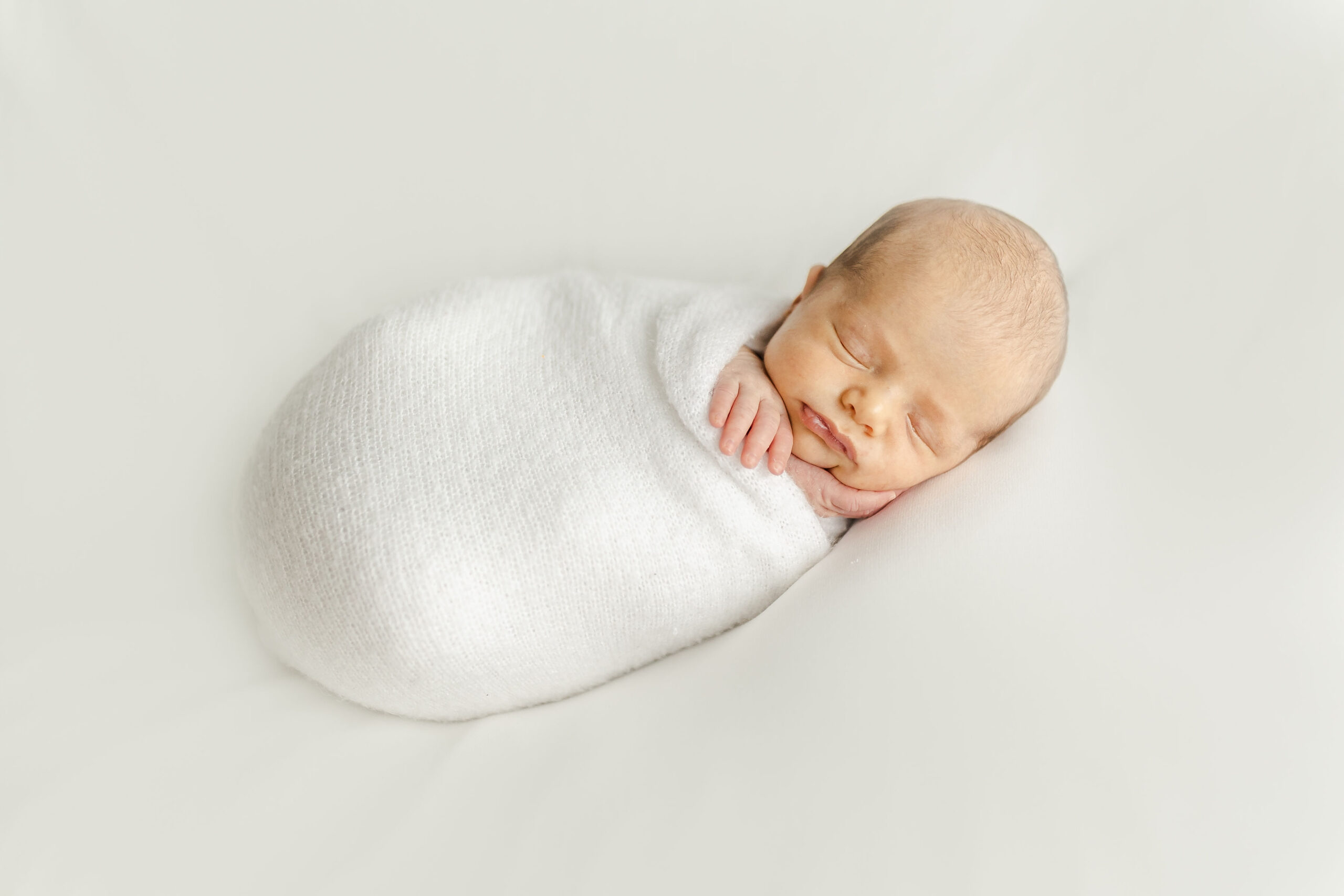 A newborn baby sleeps on its side while swaddled tightly and resting cheek on a hand babycottons miami