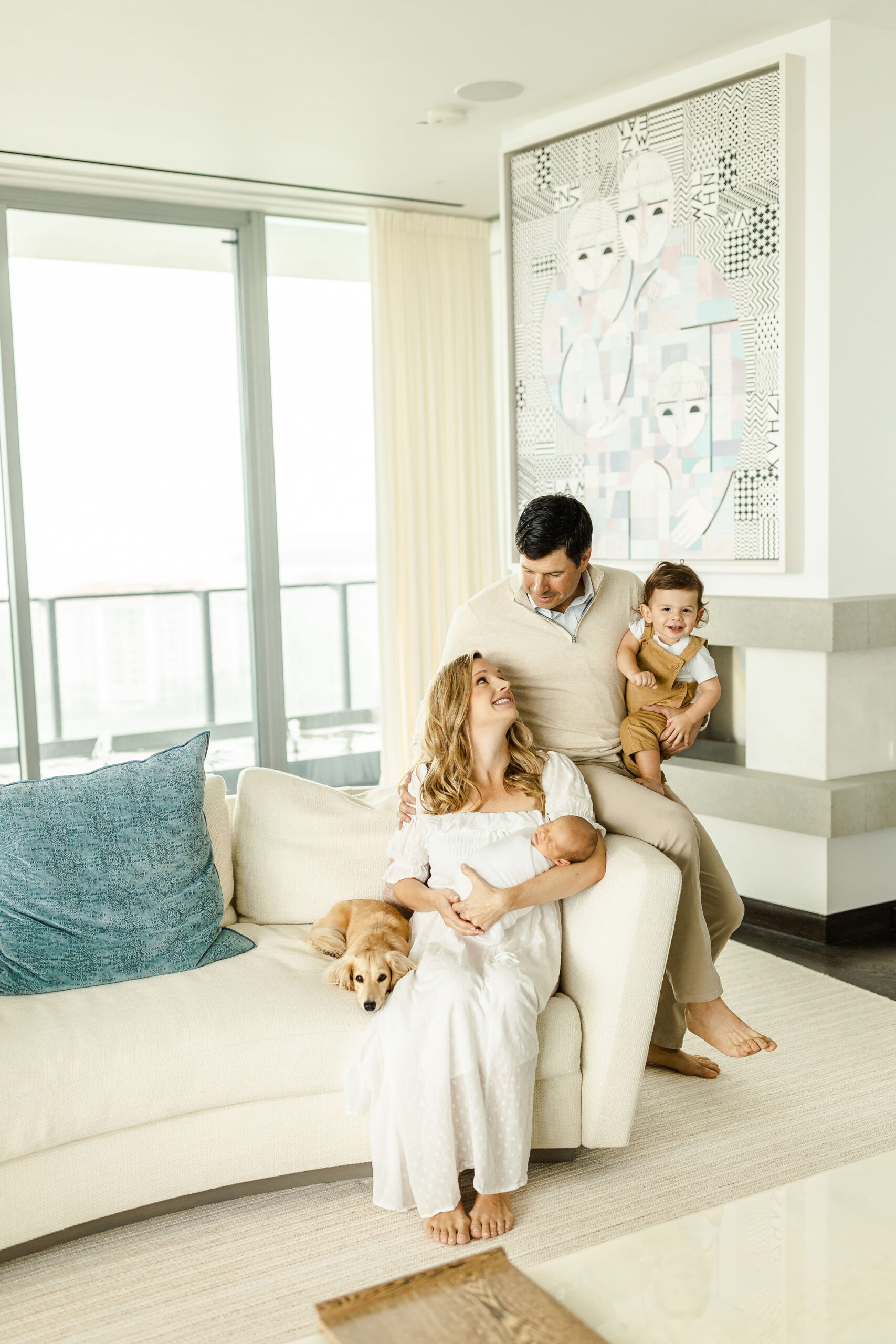 A dad sits on the edge of a couch while holding a toddler while looking down at mom and a newborn sitting on the couch with a puppy snuggle along side her babycottons miami