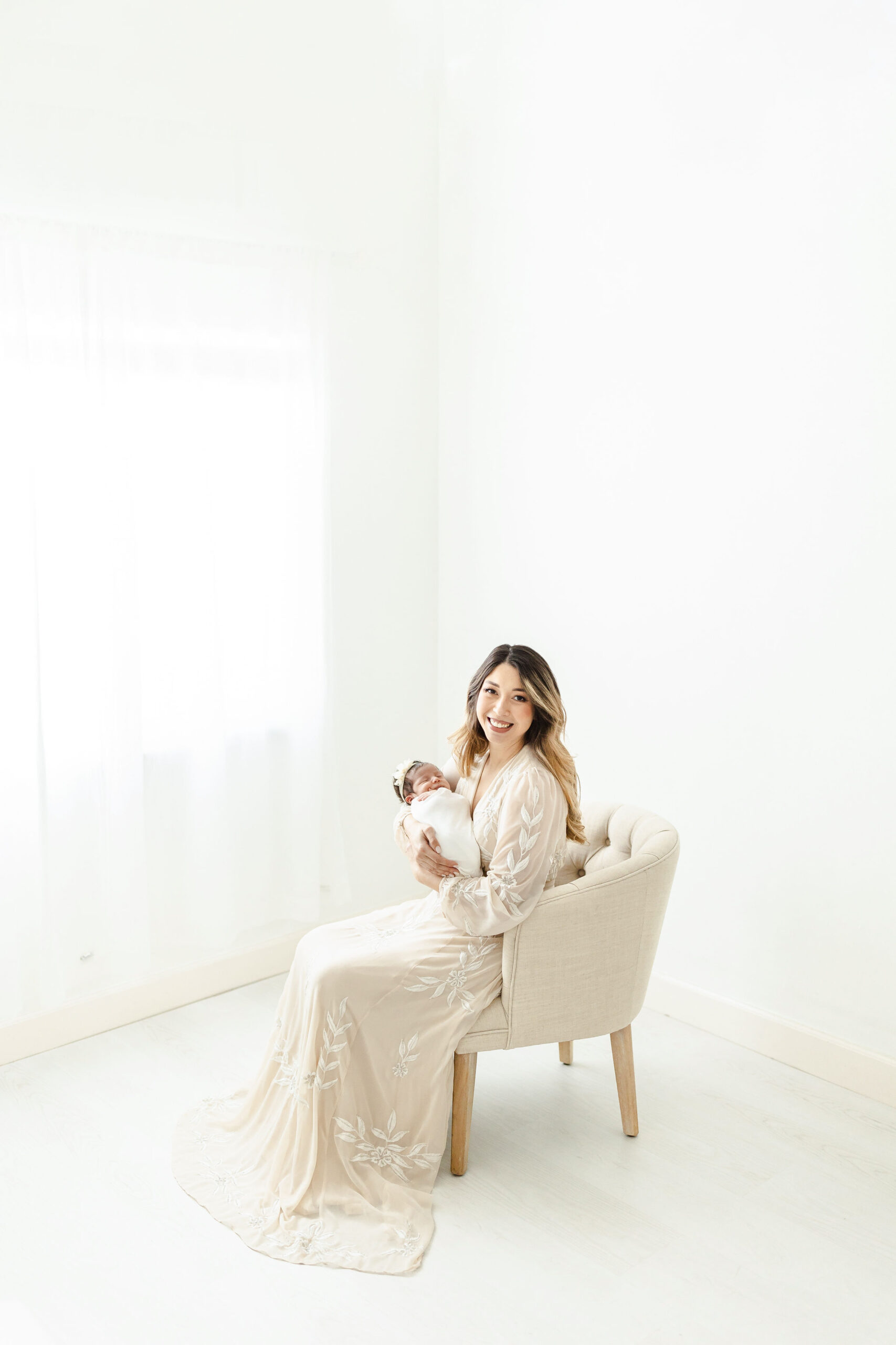 A mother to be sits in the corner of a room in front of a window on a beige chair while holding her newborn daughter
