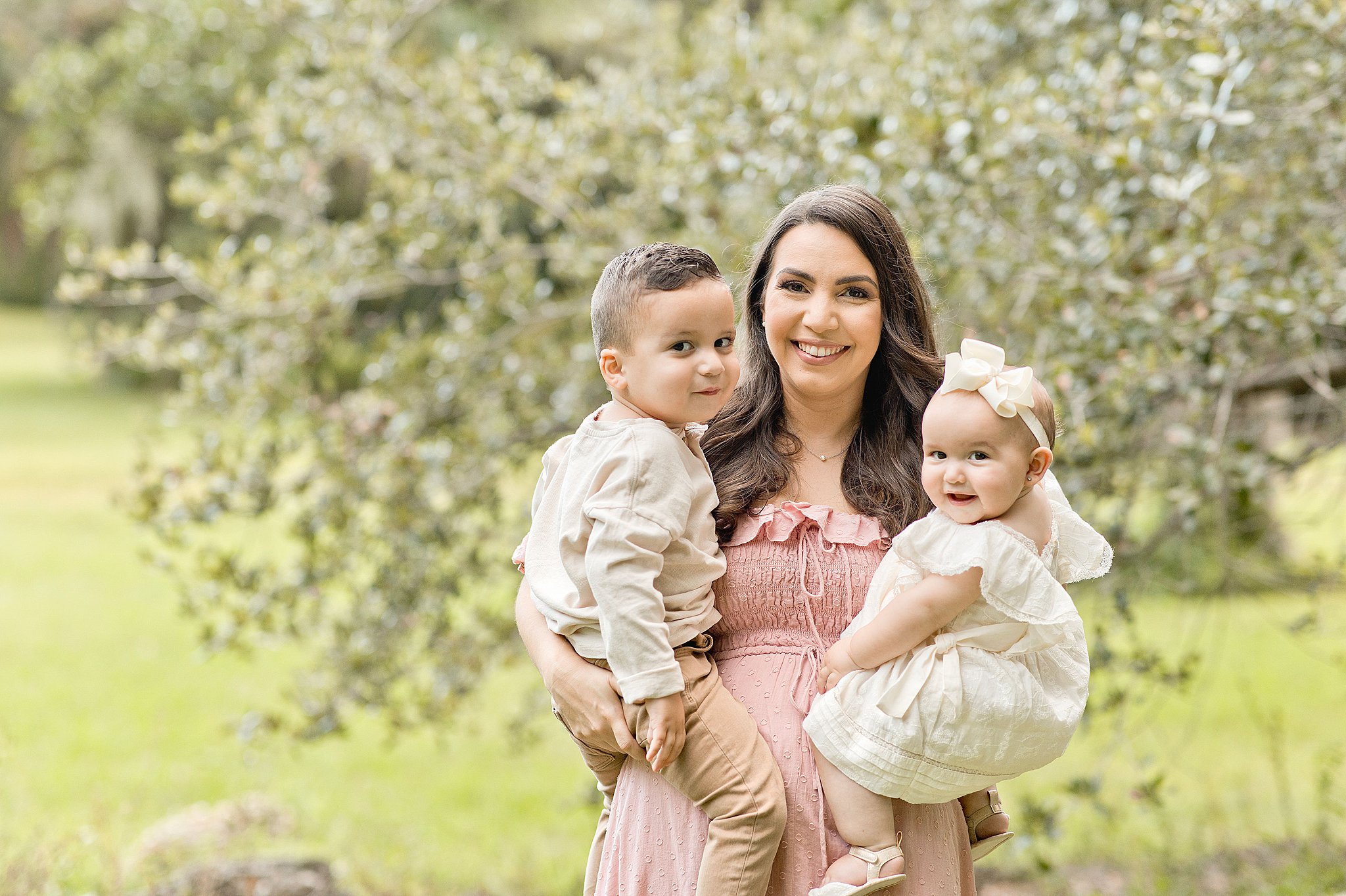 Pregnant Mother stands in a grassy field wearing a pink dress holds her two toddlers wearing khaki and tan outfits bundle of fun miami