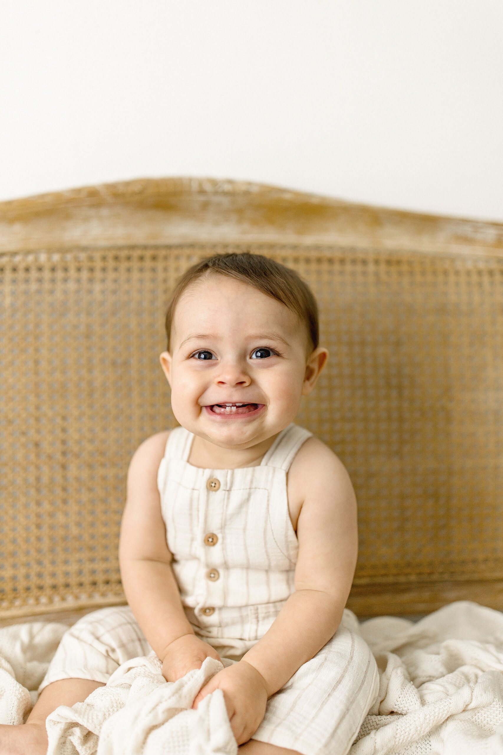 A toddler boy sits in tan overalls on a bed with a wicker headboard smiling miami toy stores