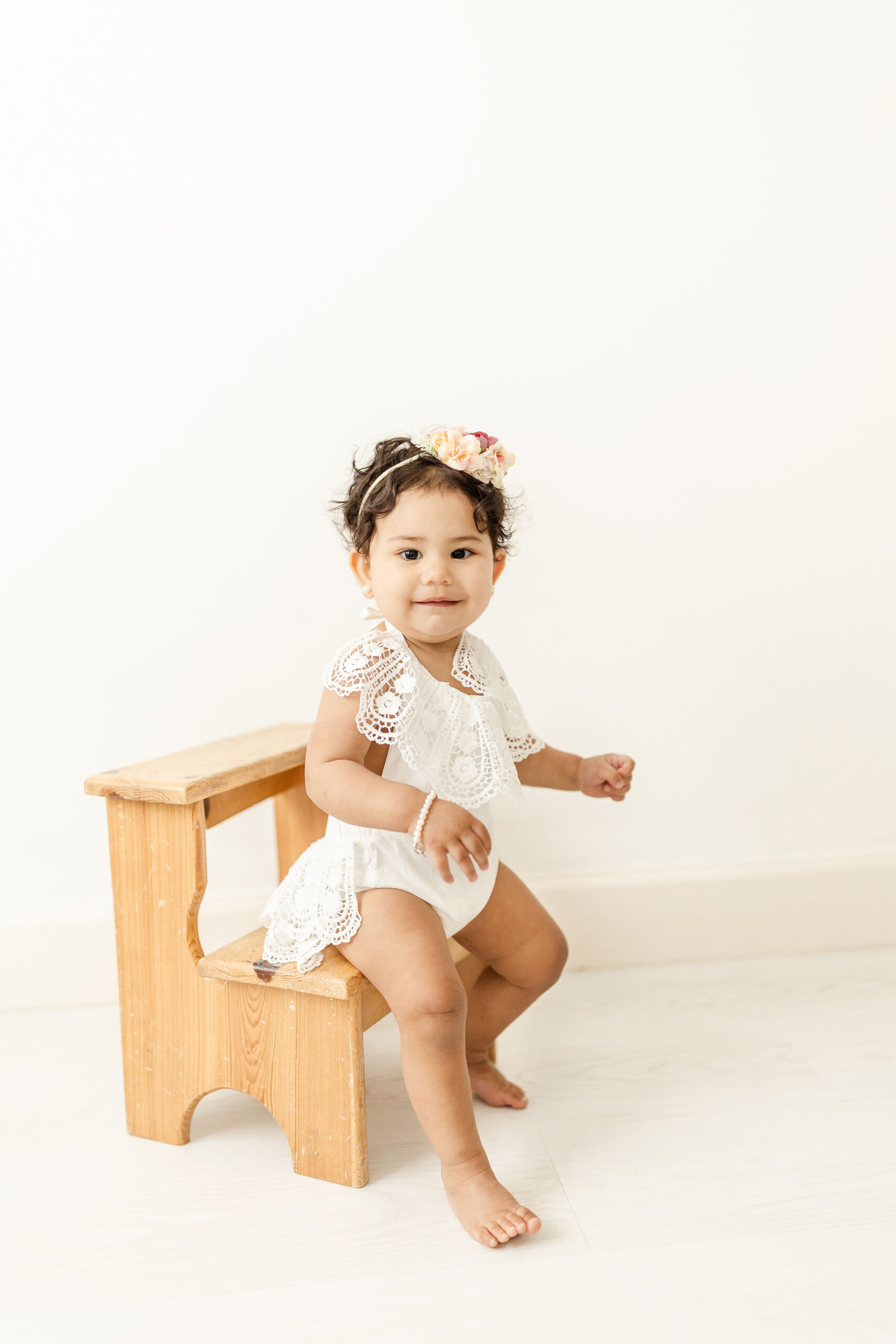 A young toddler girl sits on a wooden step stool in a studio wearing a white onesie with a large white lace collar and skirt boy meets girl miami