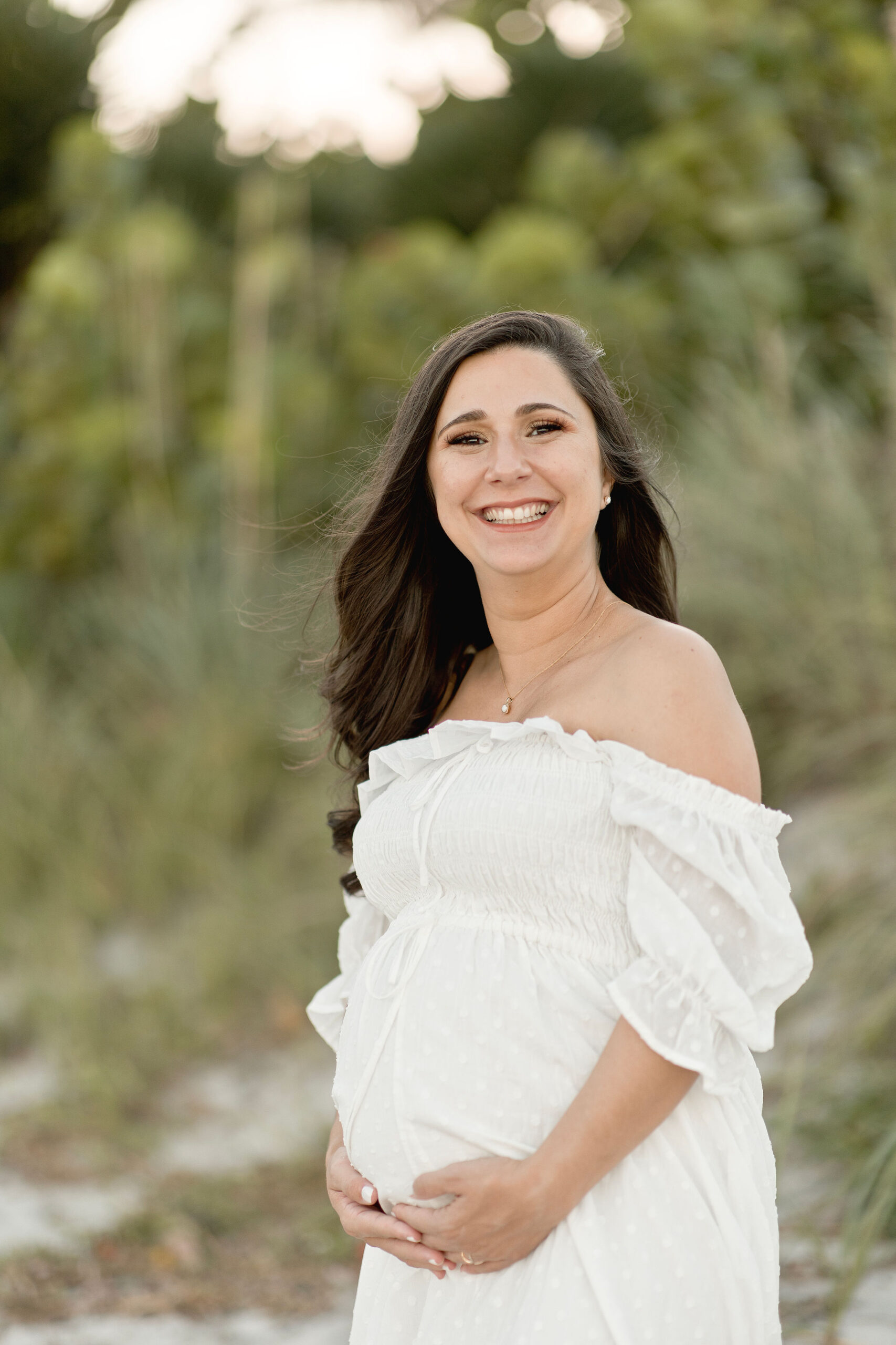 A mom to be stands in the dunes on a beach holding her bump in a white off the shoulder maternity dress maternity options of miami