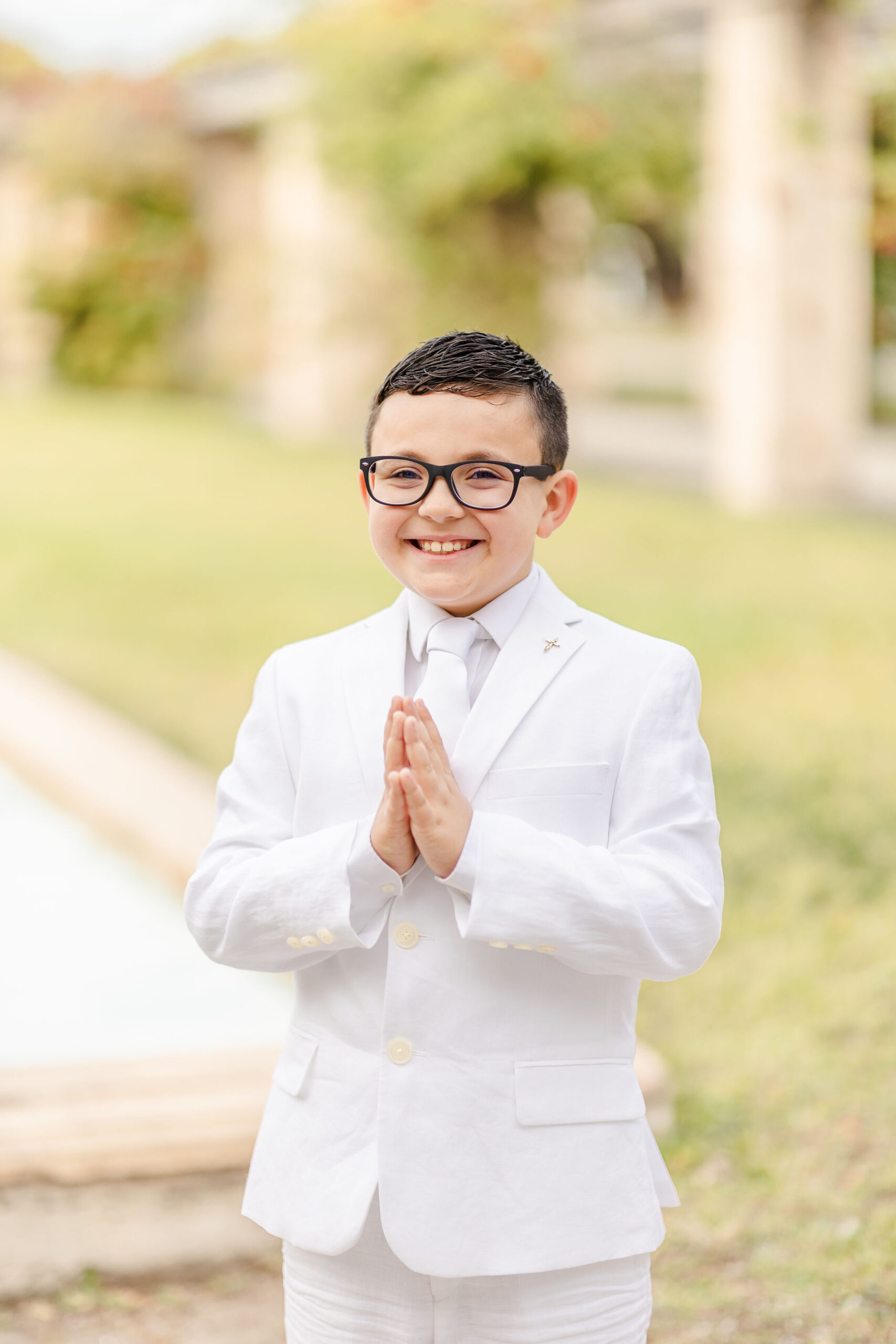 A boy holds his hands in prayer while wearing an all white suit with black glasses first communion stores in miami