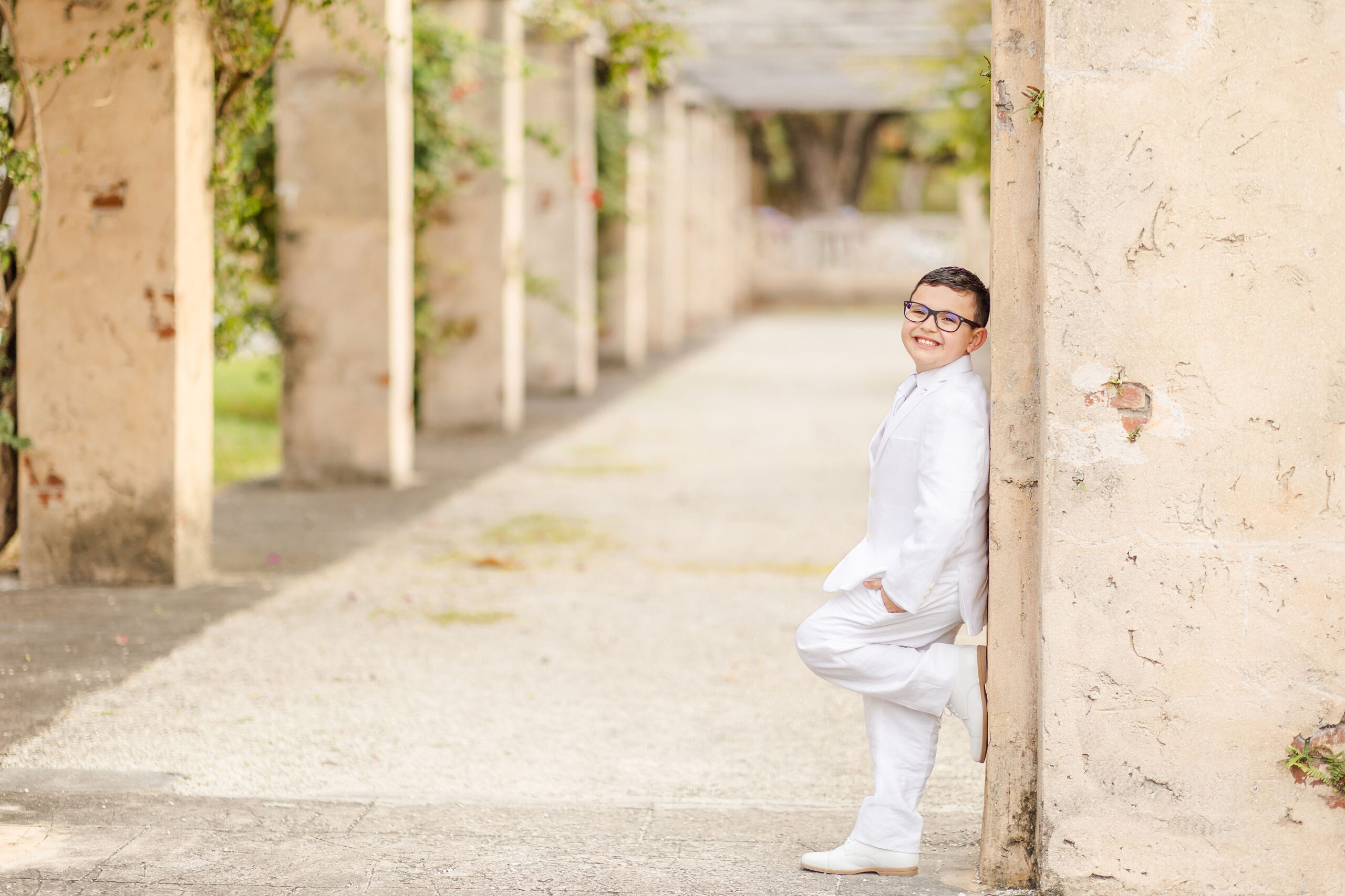 A boy in a full white suit leans against an old square column first communion stores in miami