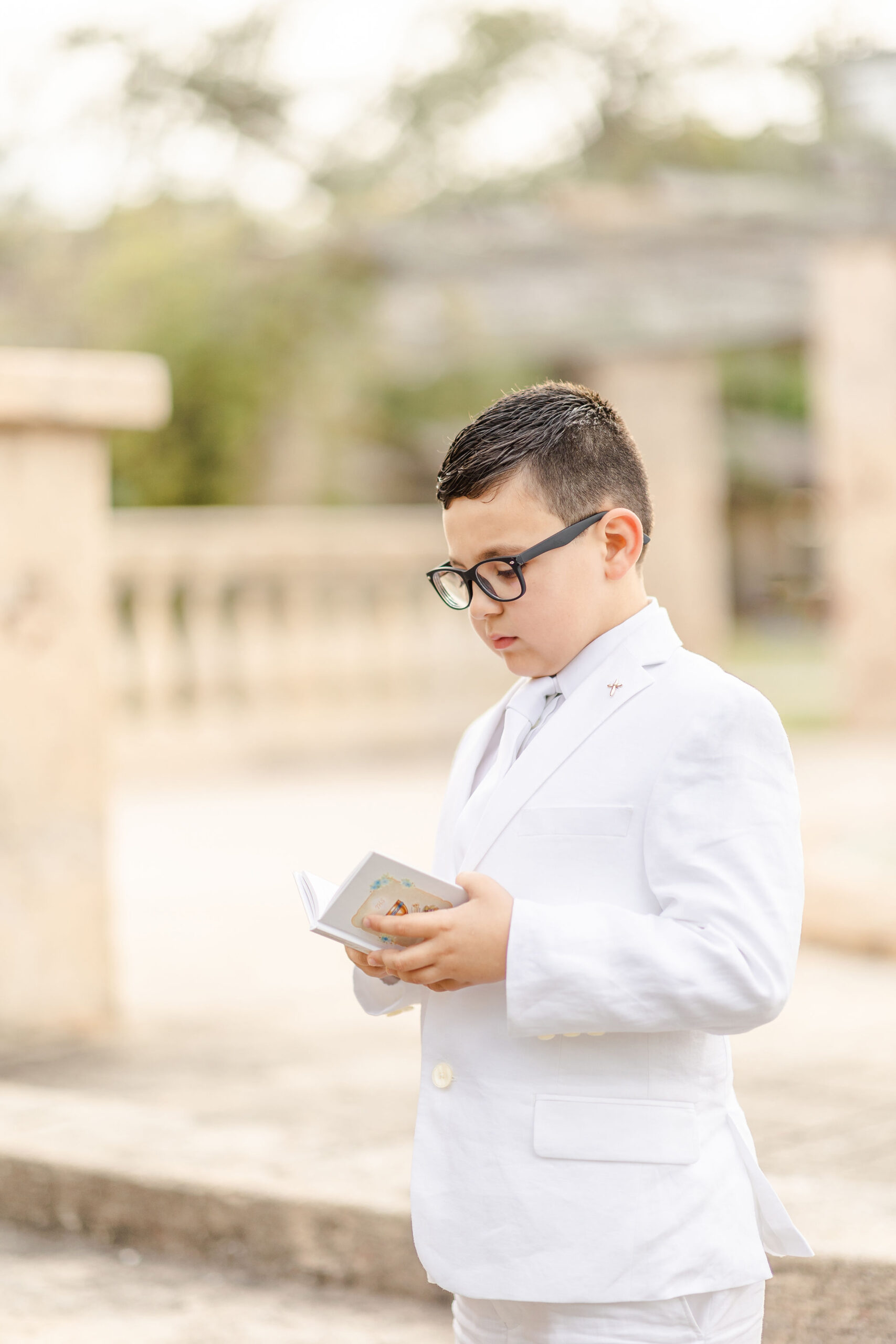 A boy wearing a white suit quietly reads his first communion prayer book on a stone path first communion stores in miami