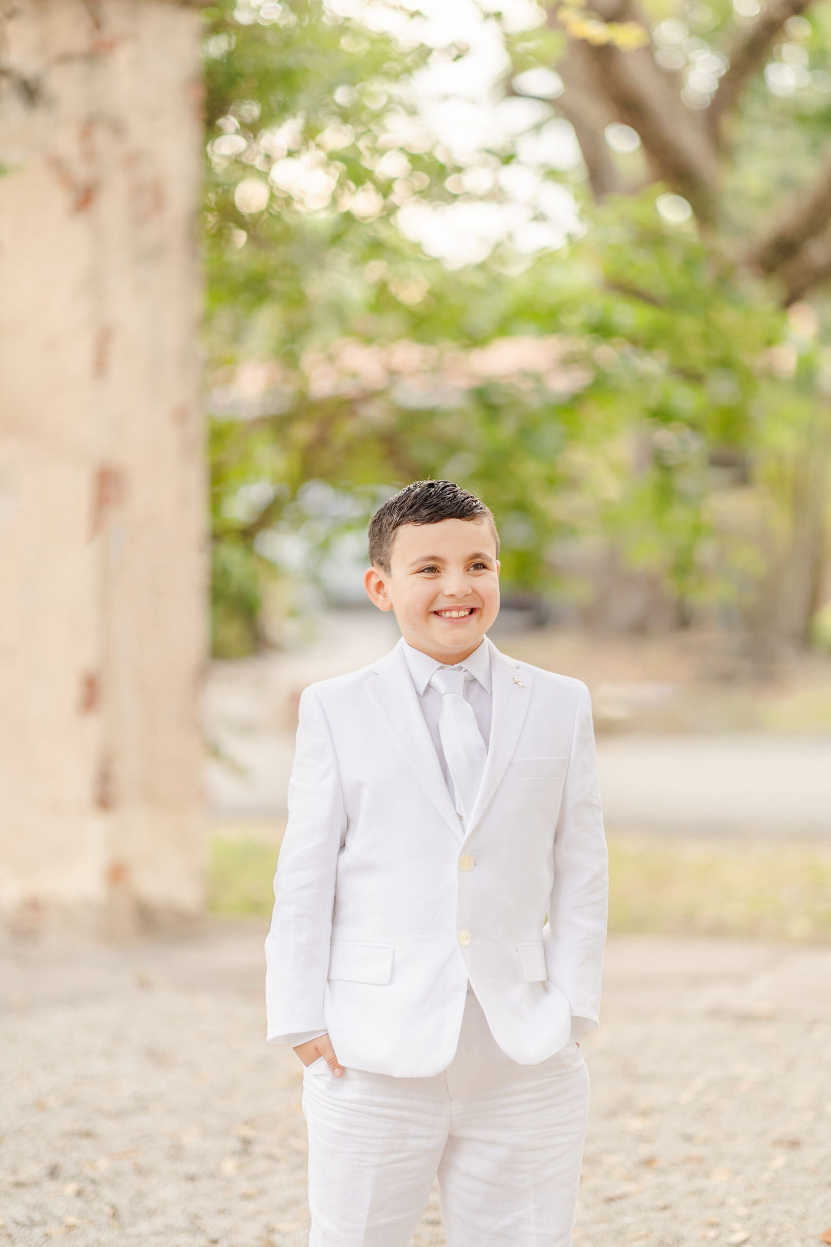 A young boy in a white suit stands with his hands in his pockets on a gravel path