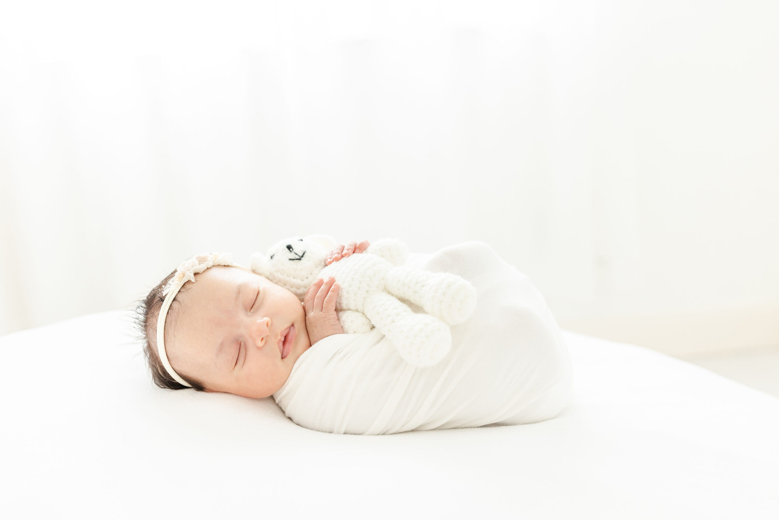 A newborn baby cuddles with a knit teddy bear while sleeping in a white swaddle on a bed under the window