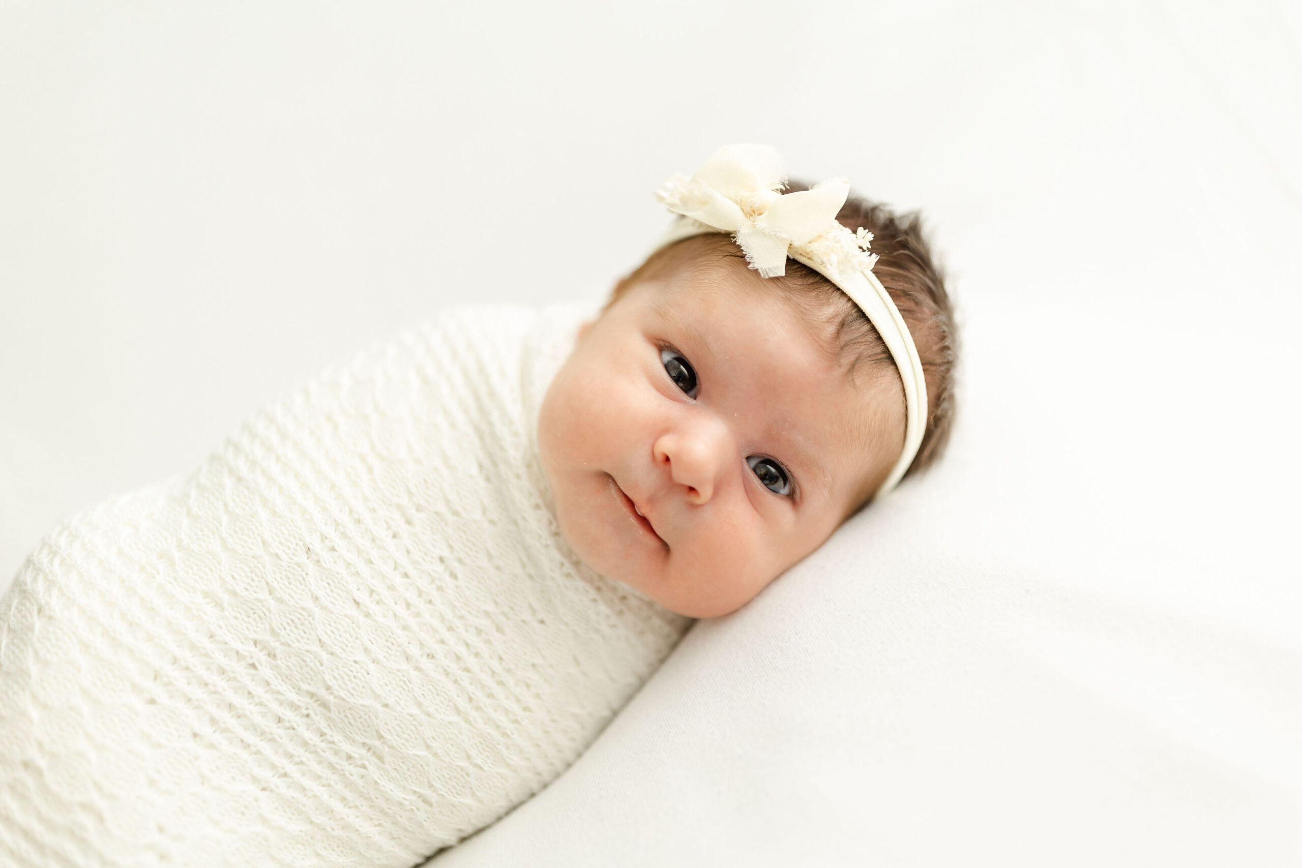 A newborn baby lays in a white lace swaddle and a white bow with eyes wide open