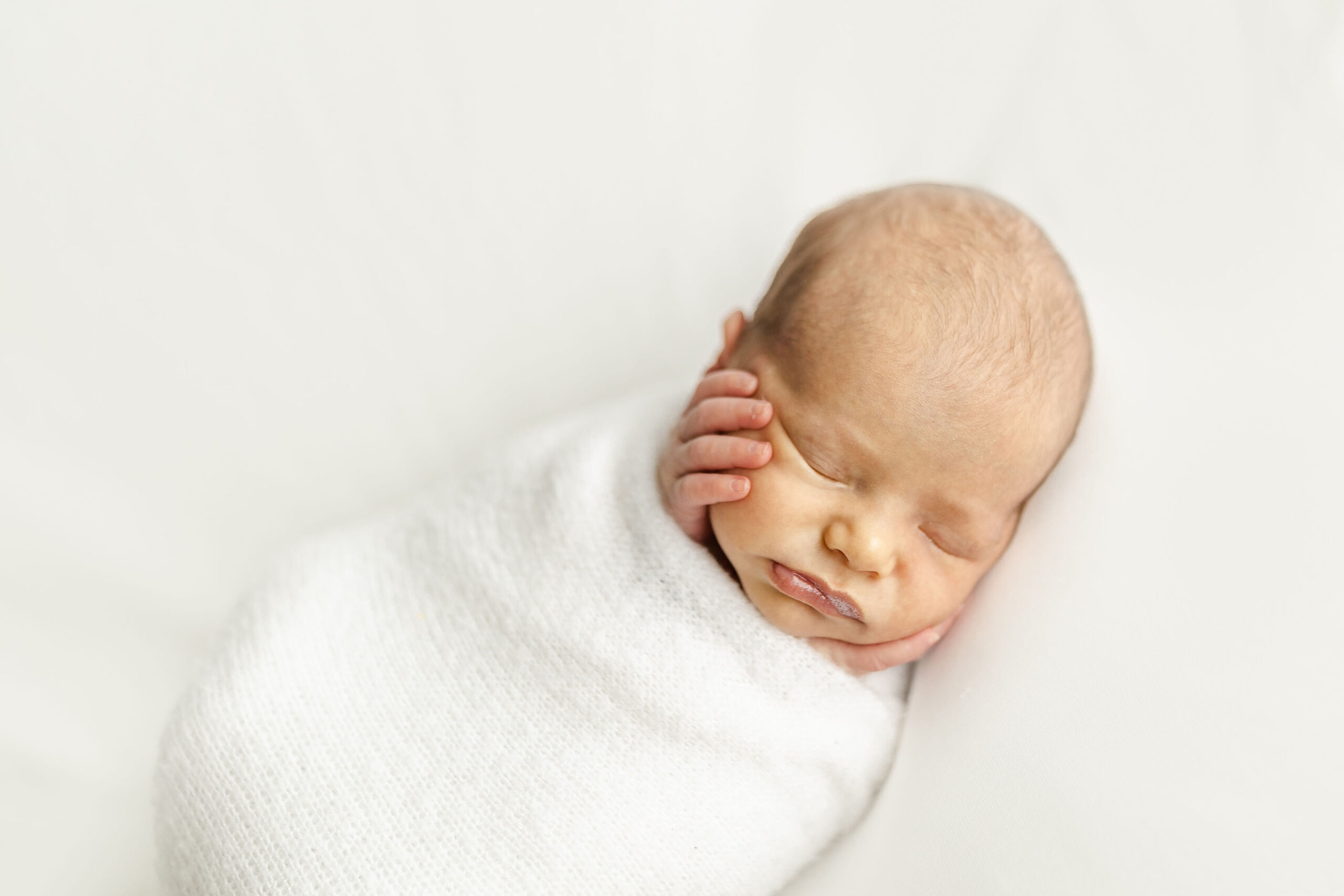 A newborn baby sleeps in a white swaddle with hands sticking out on its cheeks luna baby miami