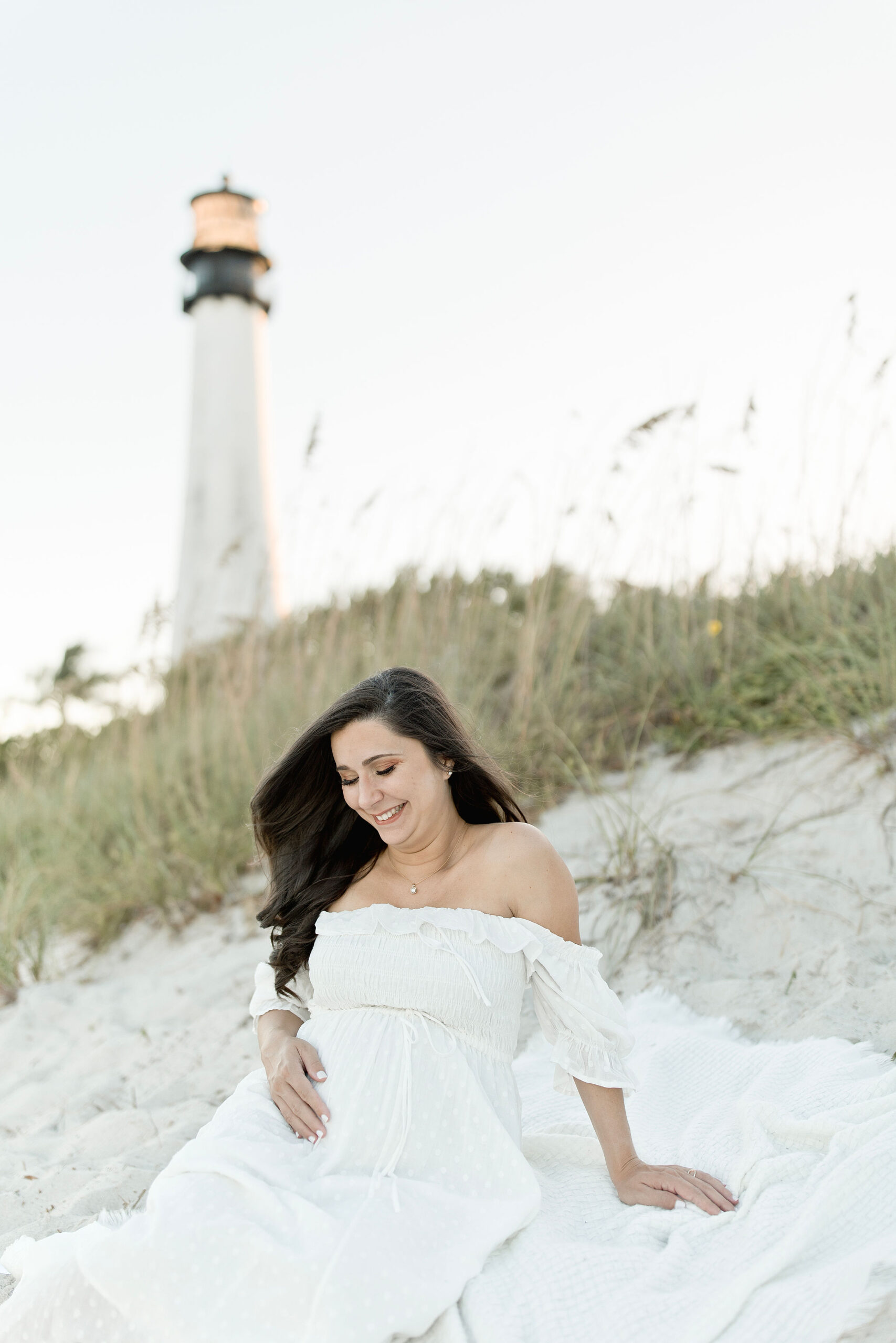 A mom to be sits on a blanket on the beach in a white maternity dress looking down at her bump miami maternity center