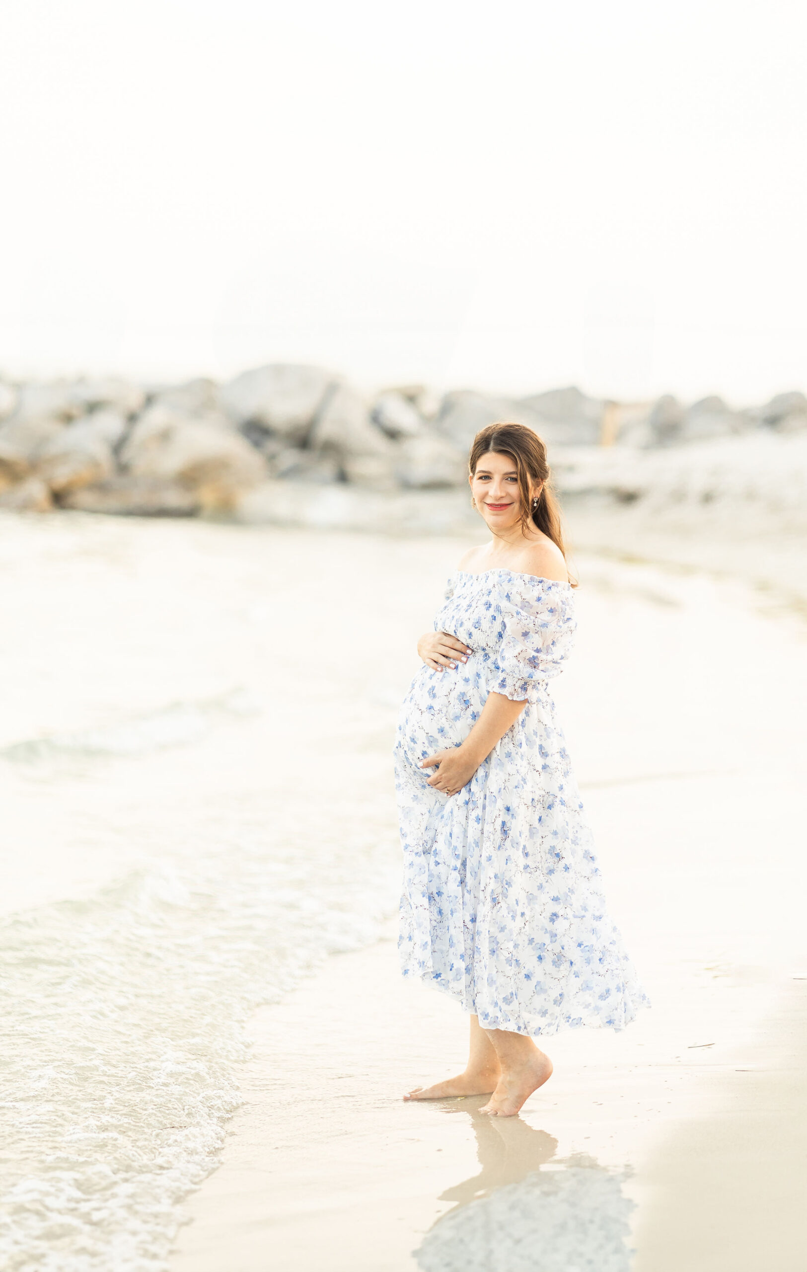 A mom to be stands on a beach barefoot in a blue flower dress with hands on her bump