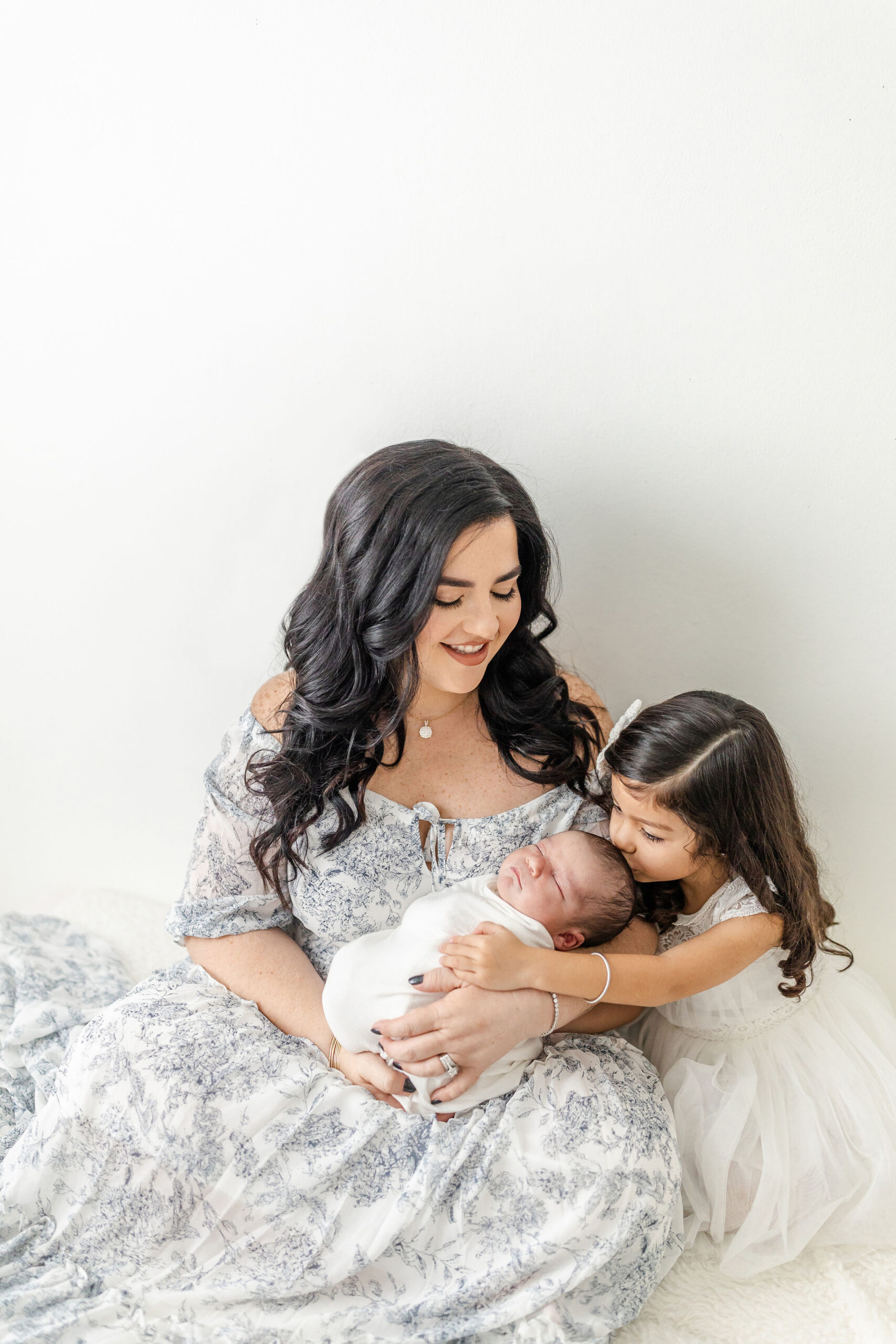 A young girl in a white dress kisses the forehead of her newborn sibling in mom's lap