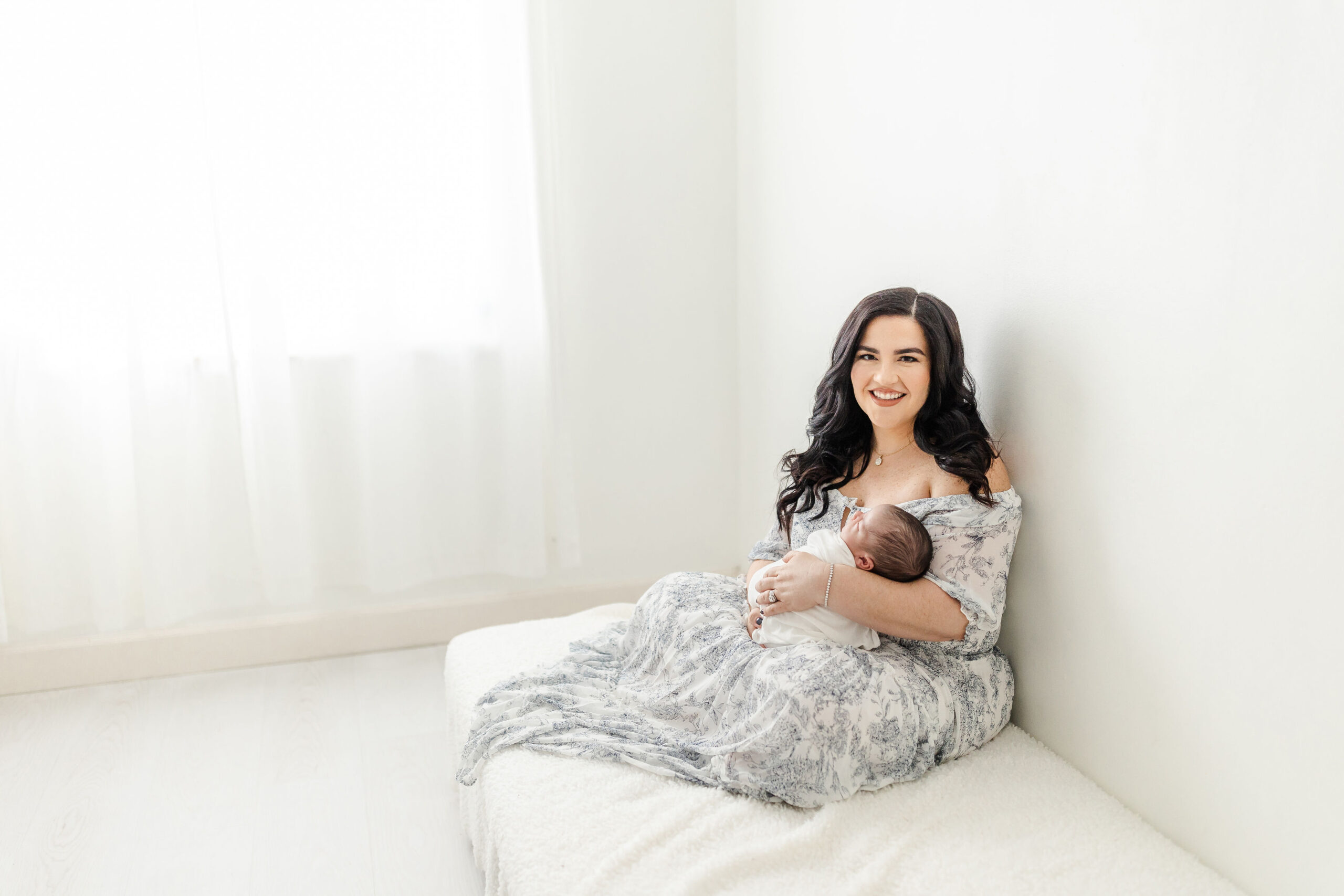 A new mom sits on a bed in a blue dress while holding her newborn baby in her lap by a window lumina massage