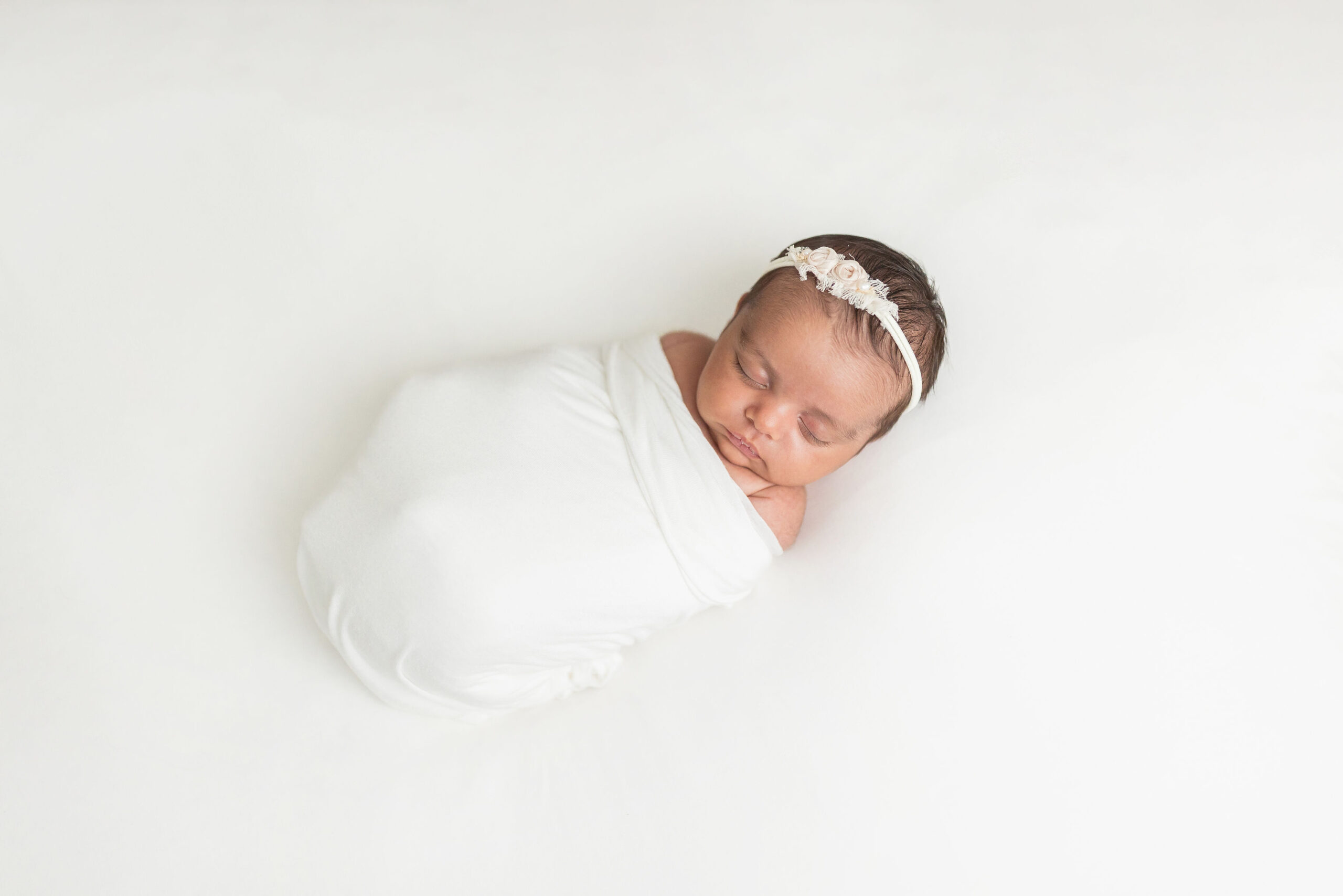 A newborn baby sleeps in a white swaddle with shoulders out on a white bed