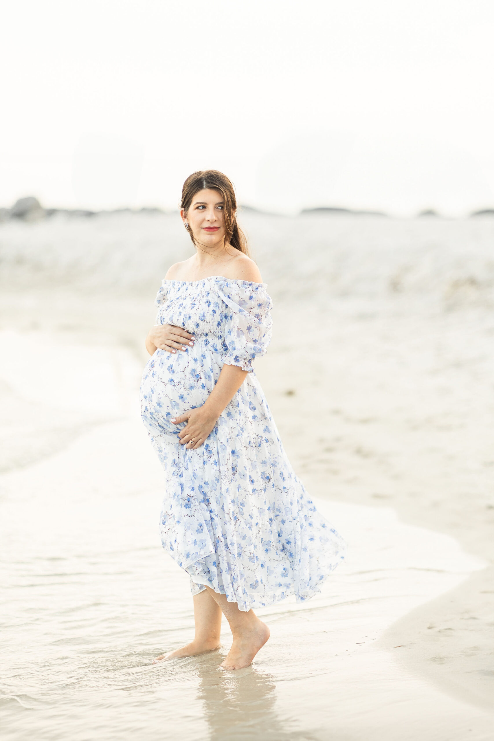 A mom to be looks over her shoulder while walking down a beach holding her bump in a blue floral dress