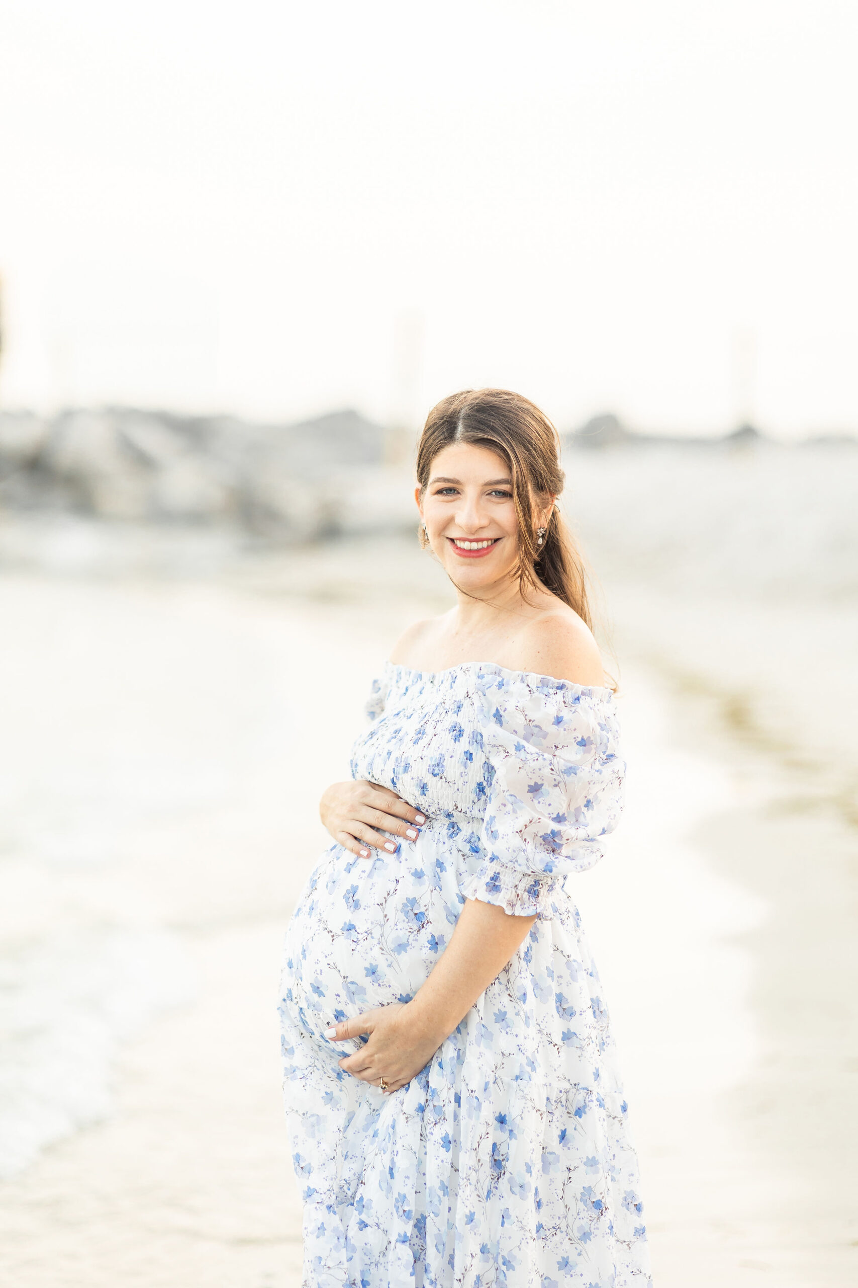 A mom to be wearing a blue floral dress stands on a beach holding her bump yoga nest