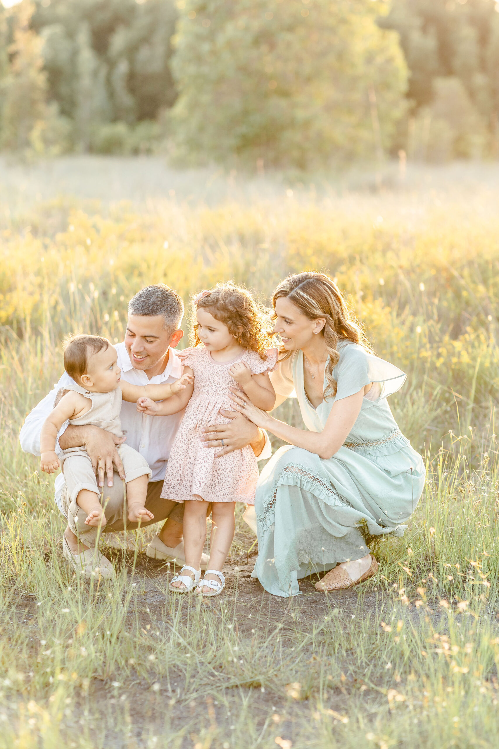 A mother and father play and explore with their toddler daughter and infant son in a field of tall grass at sunset