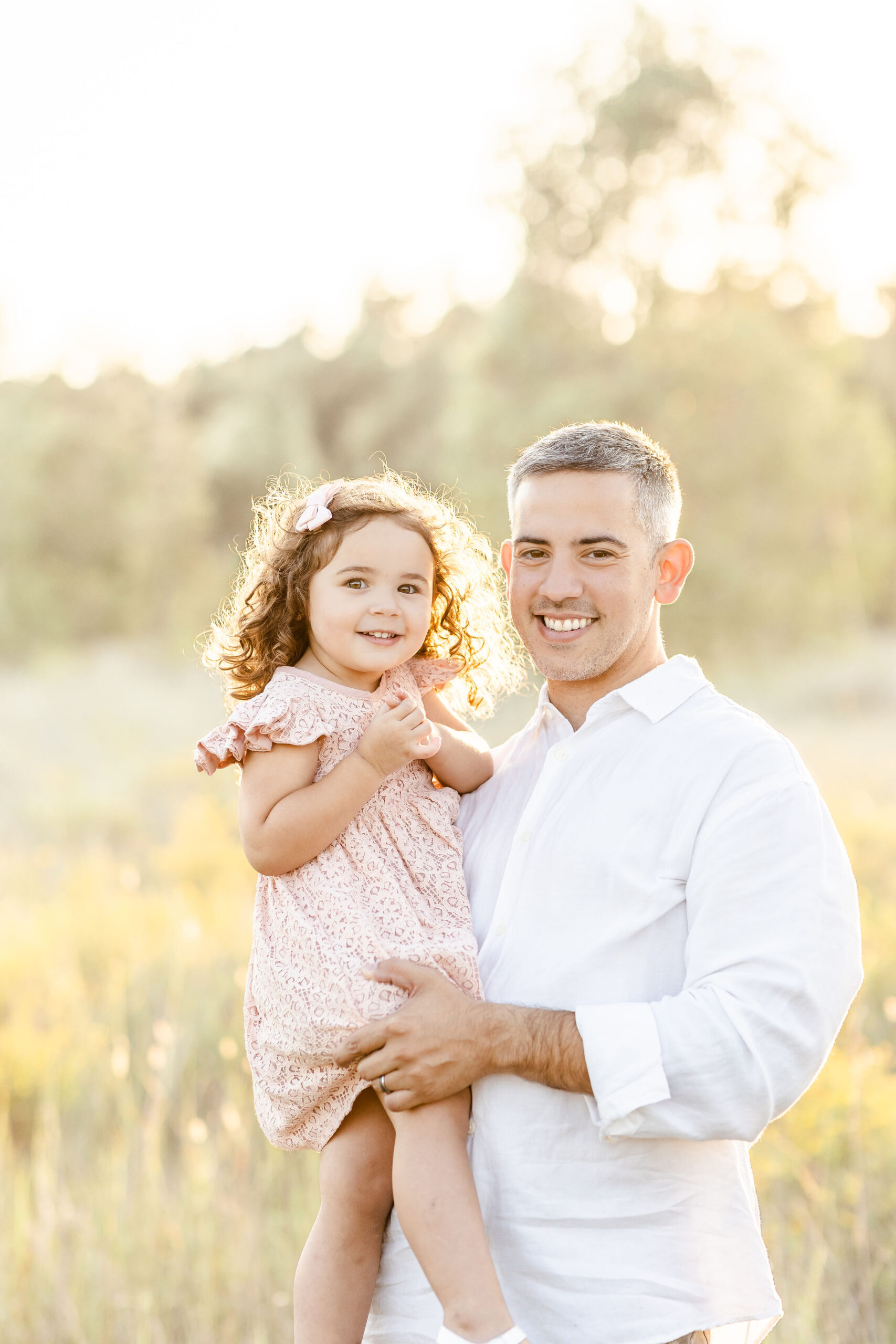 A dad in a white shirt holds his toddler daughter in a pink dress while standing in a bright grassy field at sunset ideal baby and kids