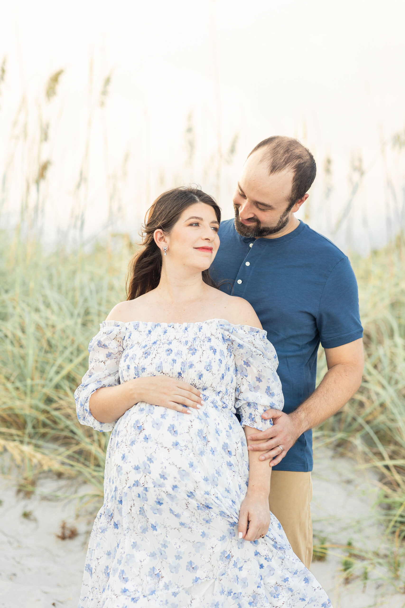 A mom to be in a blue floral dress rests her hand on her bump while looking over her shoulder to her husband behind her on a beach in the dunes