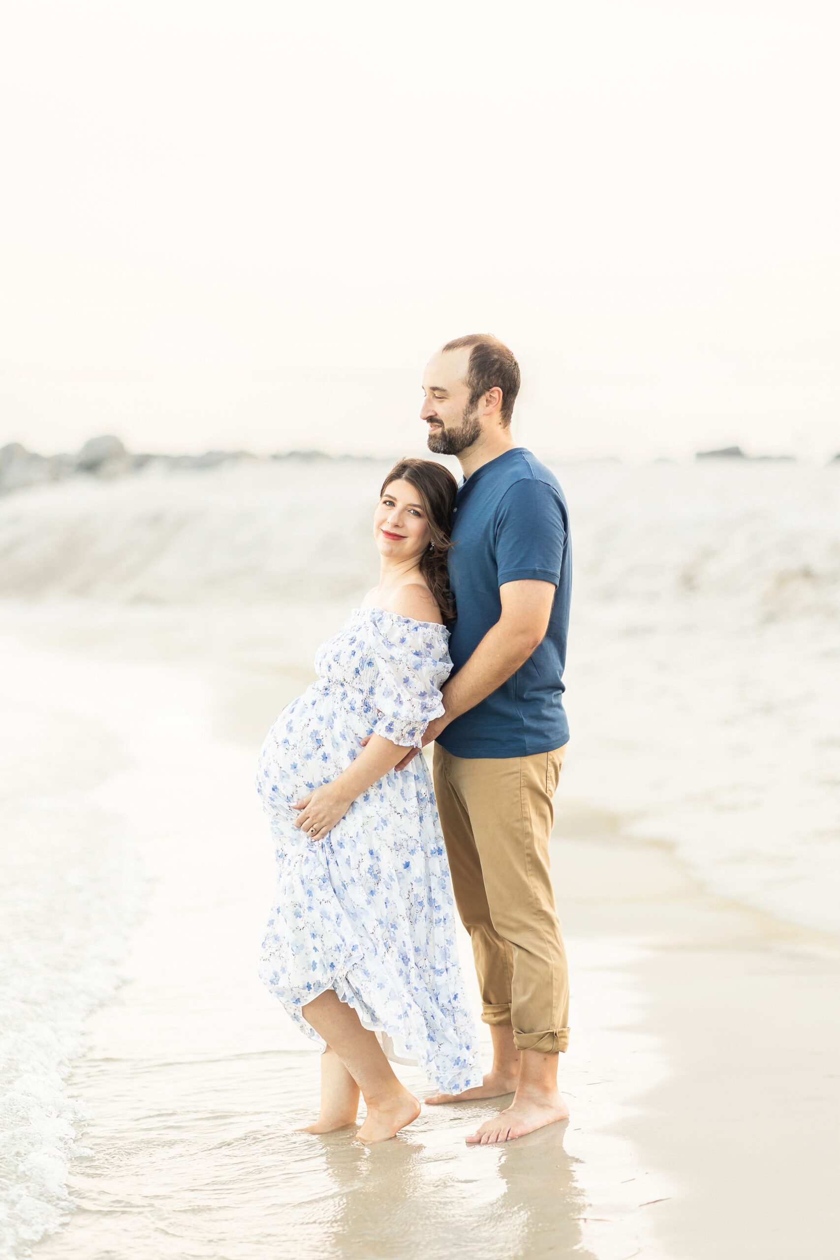A mom to be leans into her husband while standing in the water on a beach in a blue floral dress after miami prenatal massage