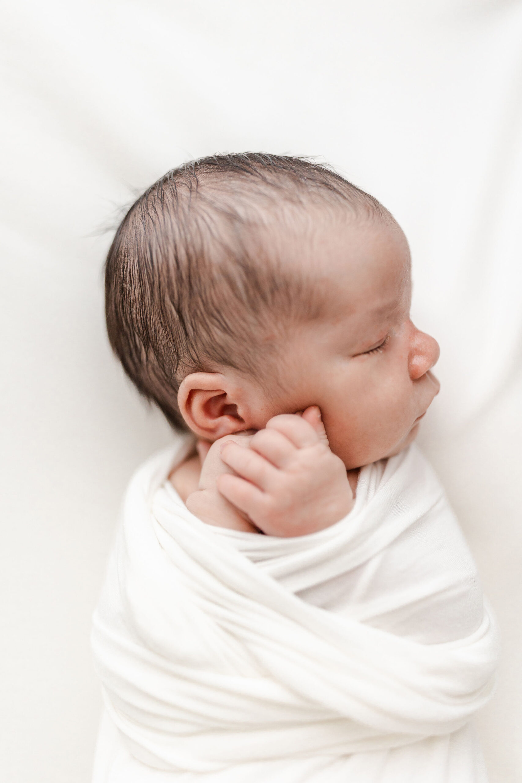 A newborn baby sleeps in a white swaddle on a white bed with hands on its cheek