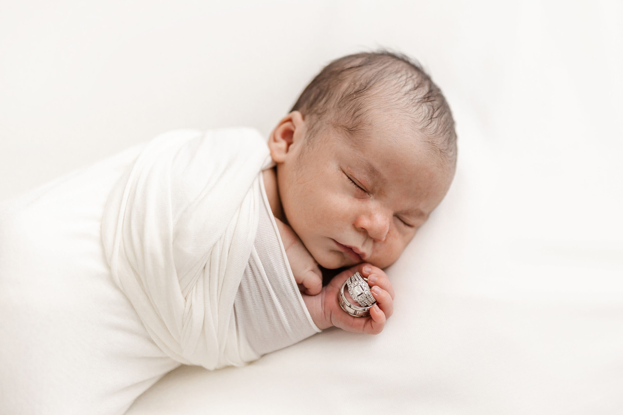 A newborn baby sleeps in a white swaddle on a bed while holding mom and dad's wedding bands spirit of life midwifery