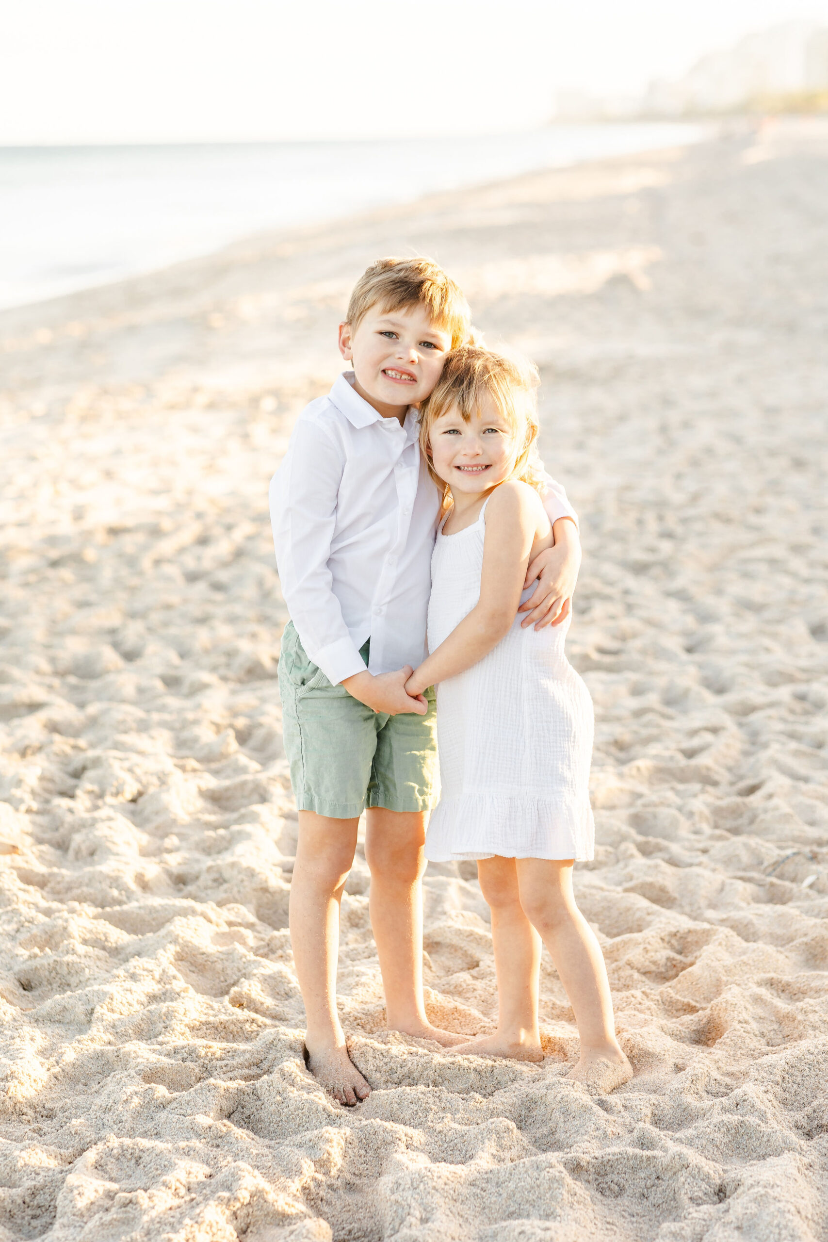 A young boy in shorts and a white button down shirt stands on a beach hugging onto his younger sister and holding her hand warm hearts children's boutique