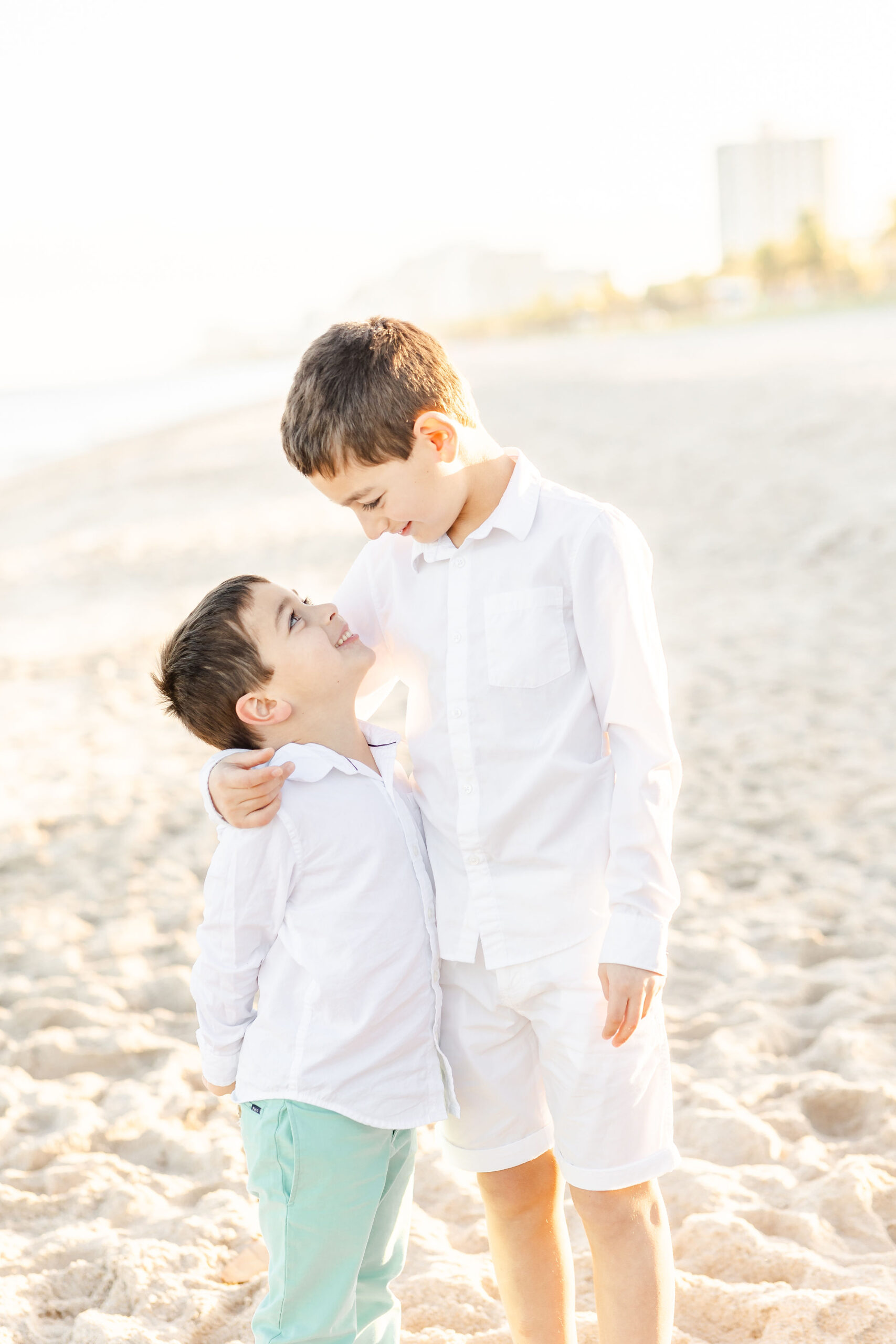 A young boy stands on a beach looking down at his younger brother with his arm around him warm hearts children's boutique