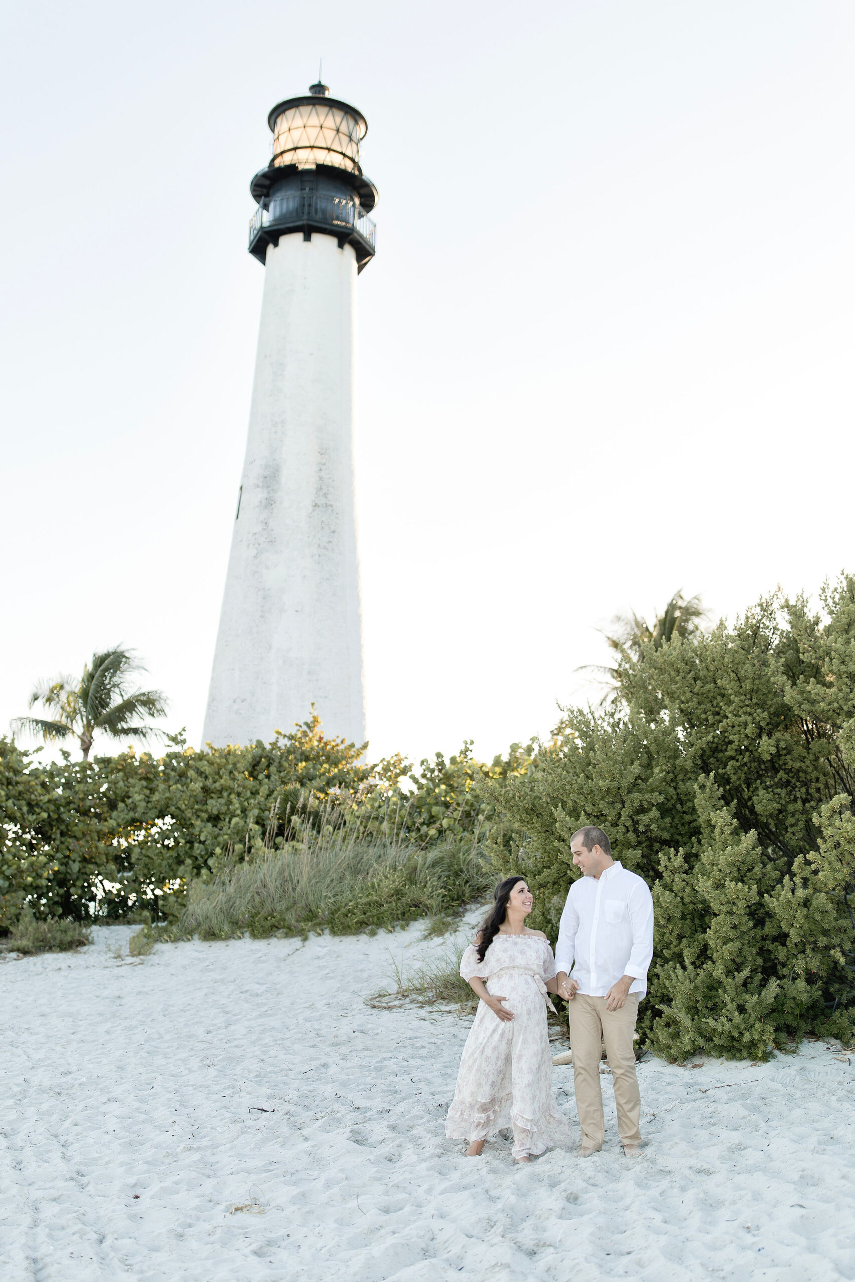A mom to be in a white maternity dress walks through the dunes holding hands with her partner in a white shirt by a lighthouse at sunset