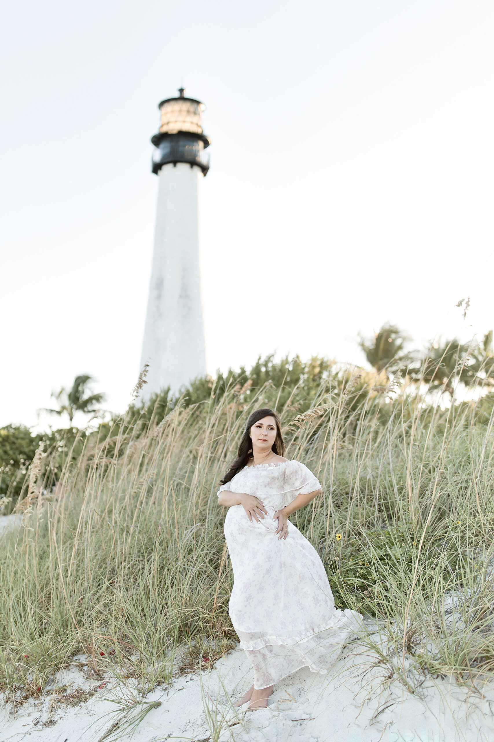 A mom to be stands on a windy beach dune by a lighthouse while holding her bump with help from maternal fetal medicine miami