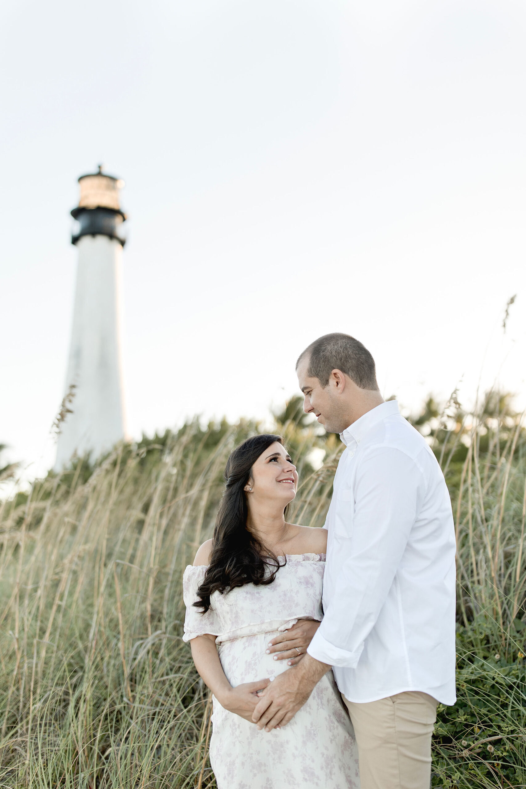 A mom to be in a white dress stands in the tall dunes with her husband in front of a lighthouse with the help of maternal fetal medicine miami