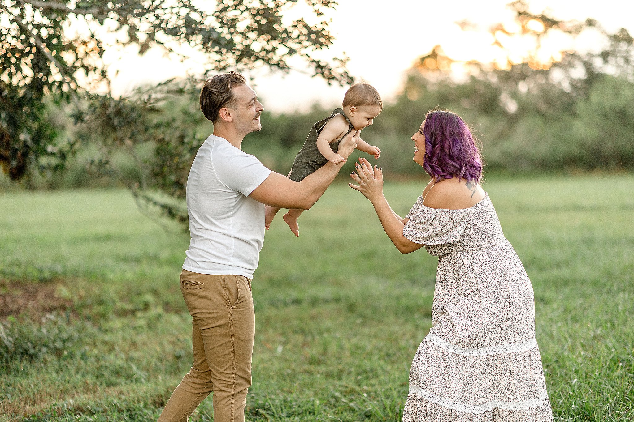 A father lifts his toddler son above mom as they stand and play in a field at sunset