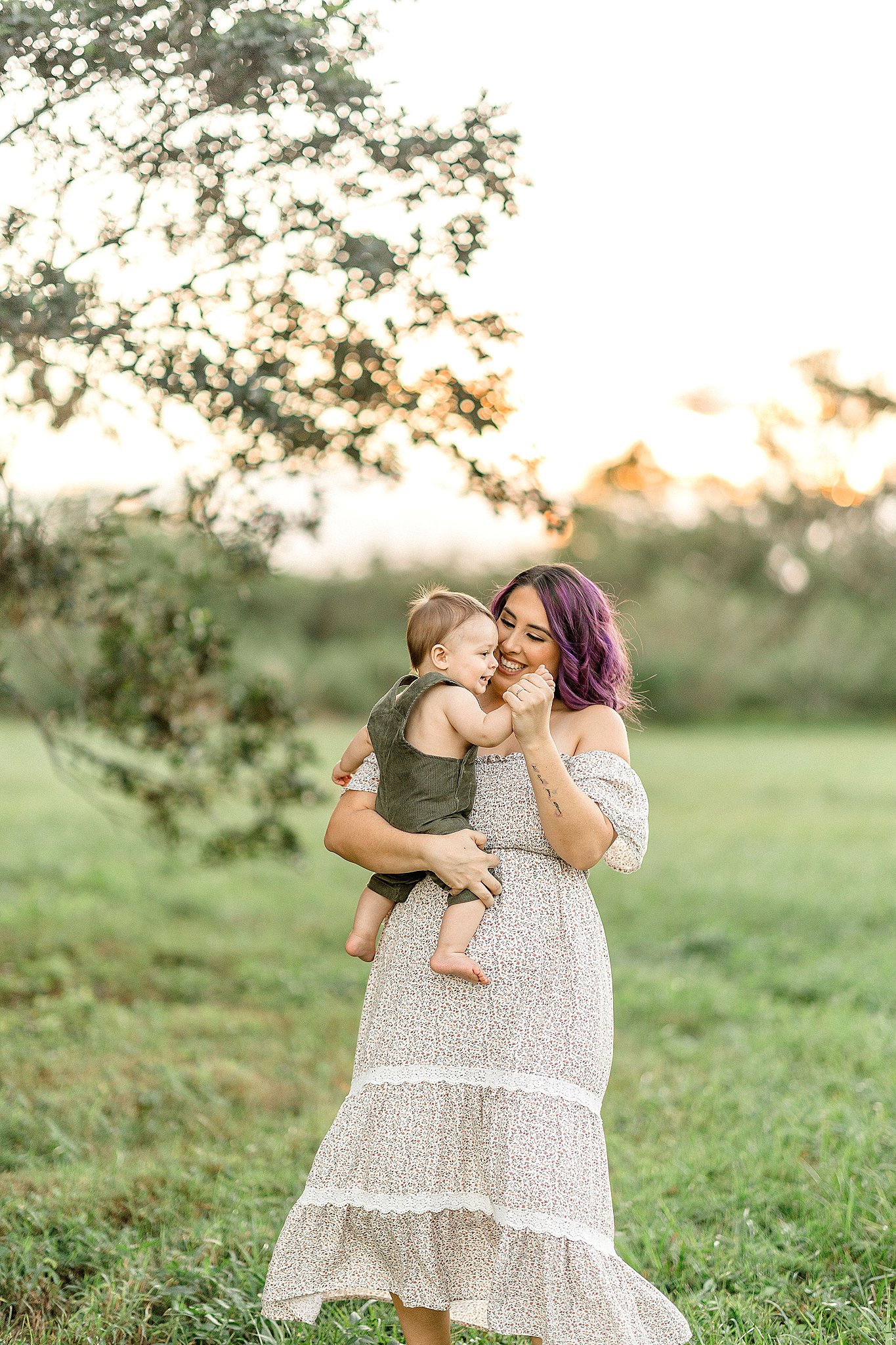 A mom with purple hair dances with her toddler son in green overalls in a grassy field at sunset after miami daycare