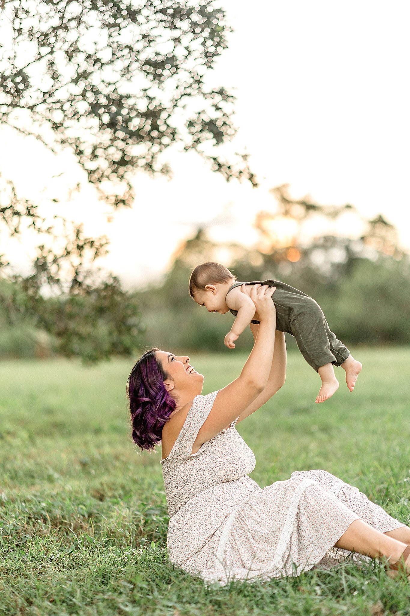 A mom in a white dress sits in the grass under a tree at sunset lifting and playing with her toddler son in green overalls after a day at a miami daycare