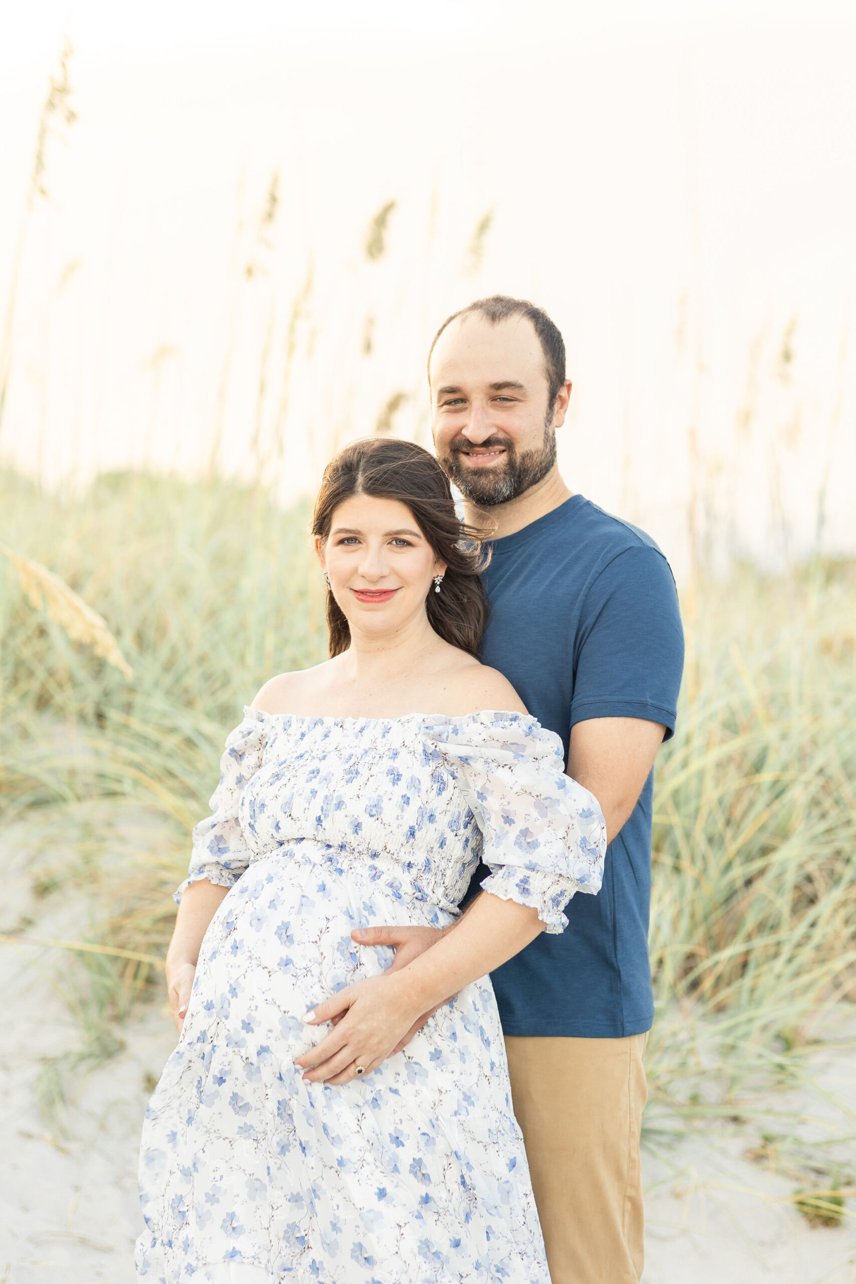 A mom to be leans into her husband as they stand in a dune on a beach with hands on the bump