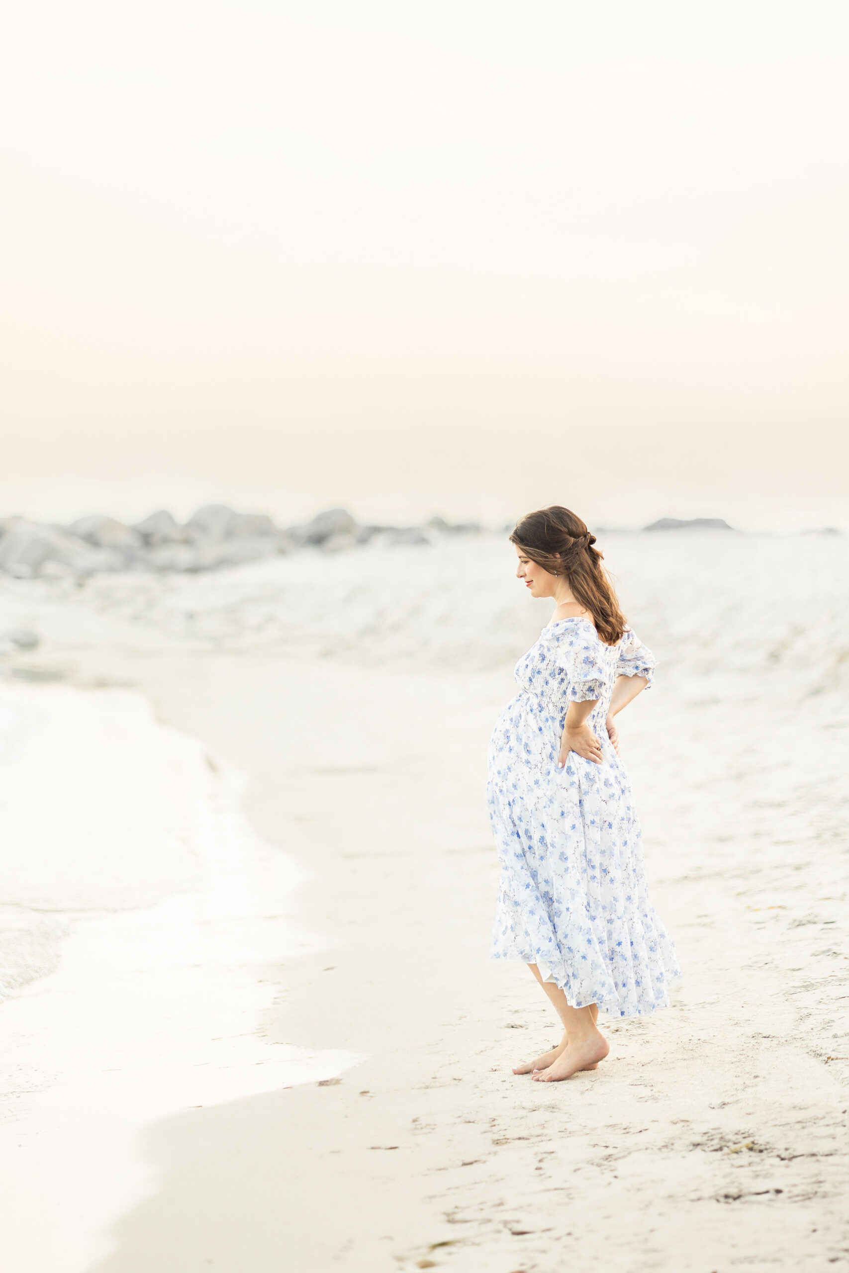 A mom to be watches the water on the beach in a blue floral dress at sunset after attending birth classes miami