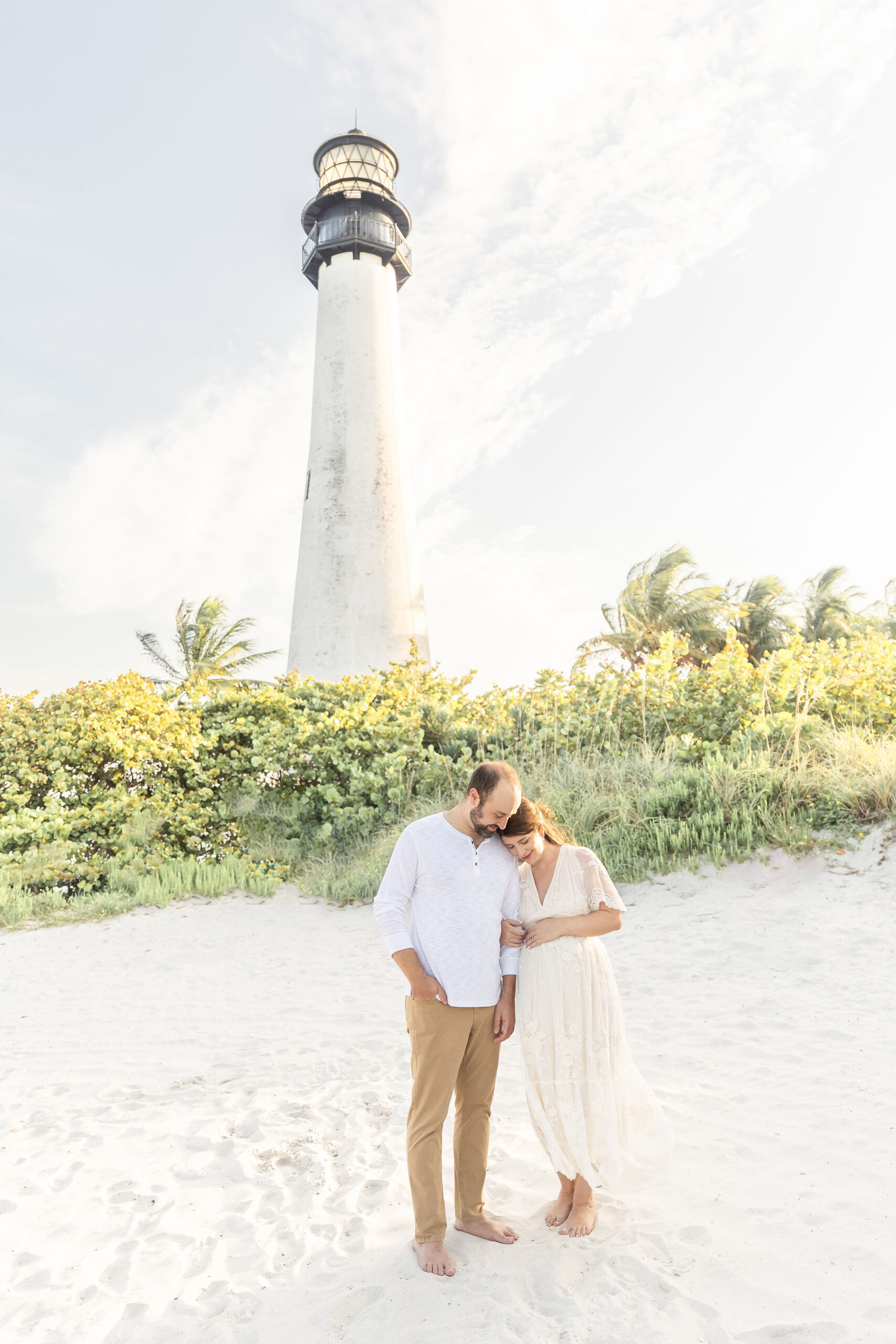 A mom to be in a beige dress leans on her husbands arm as they stand on a beach by a lighthouse after visiting a miami birthing center